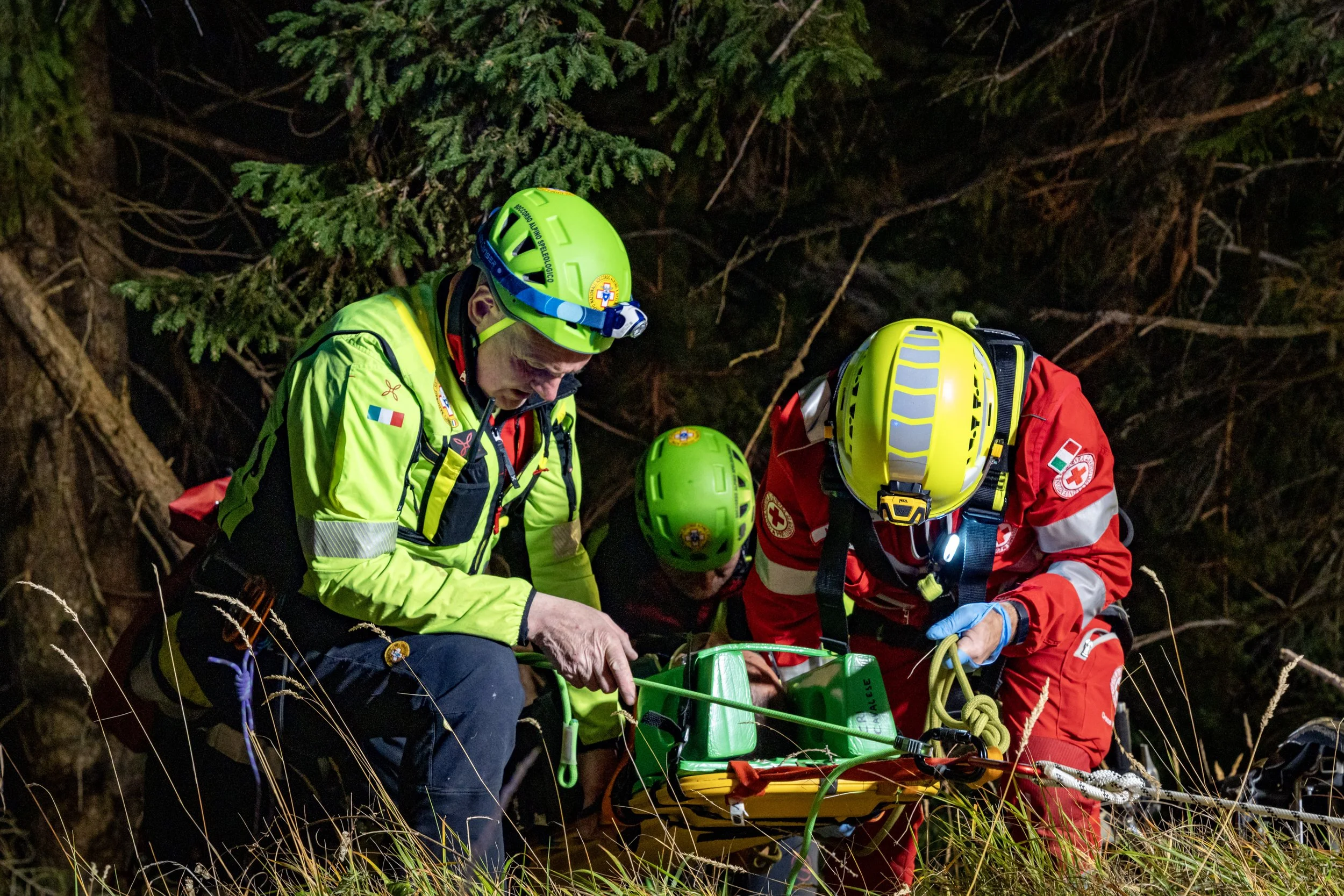 Nighttime rescue scene with emergency responders in bright uniforms and helmets, working in a dense forest to assist a person.