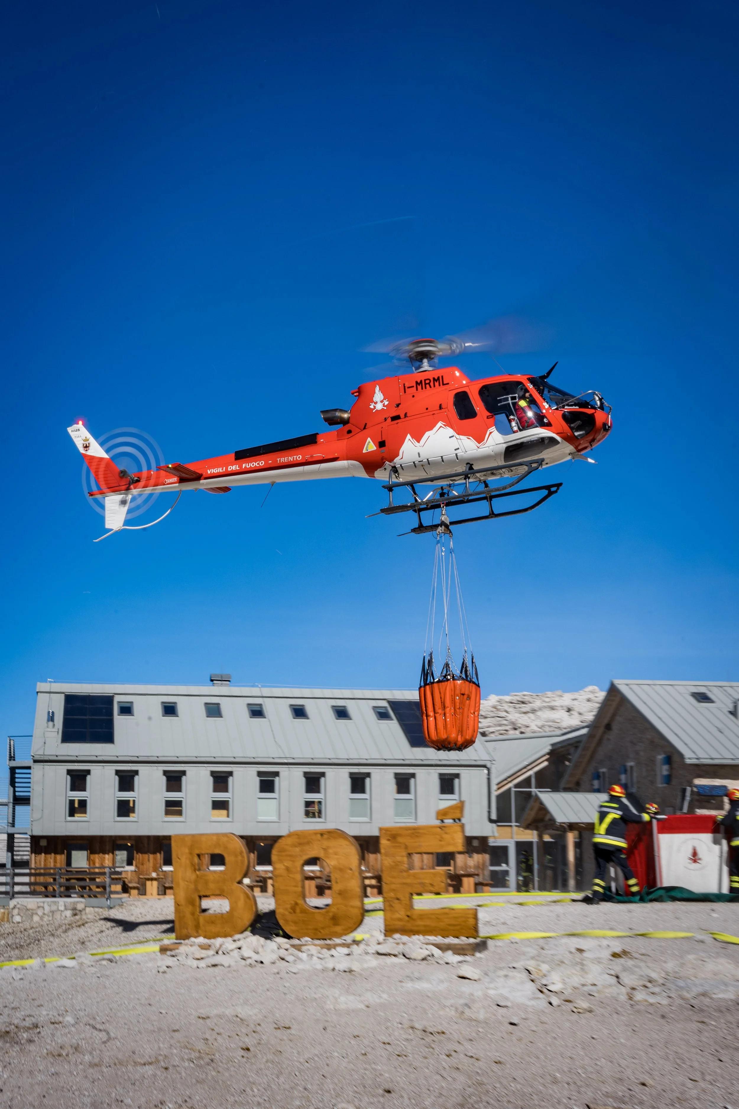 A red helicopter is flying in a clear blue sky, carrying an orange container underneath. Firefighters are on the ground wearing black and yellow gear near a wooden sign that says 'BOLZANO,' in front of a building with modern architecture.