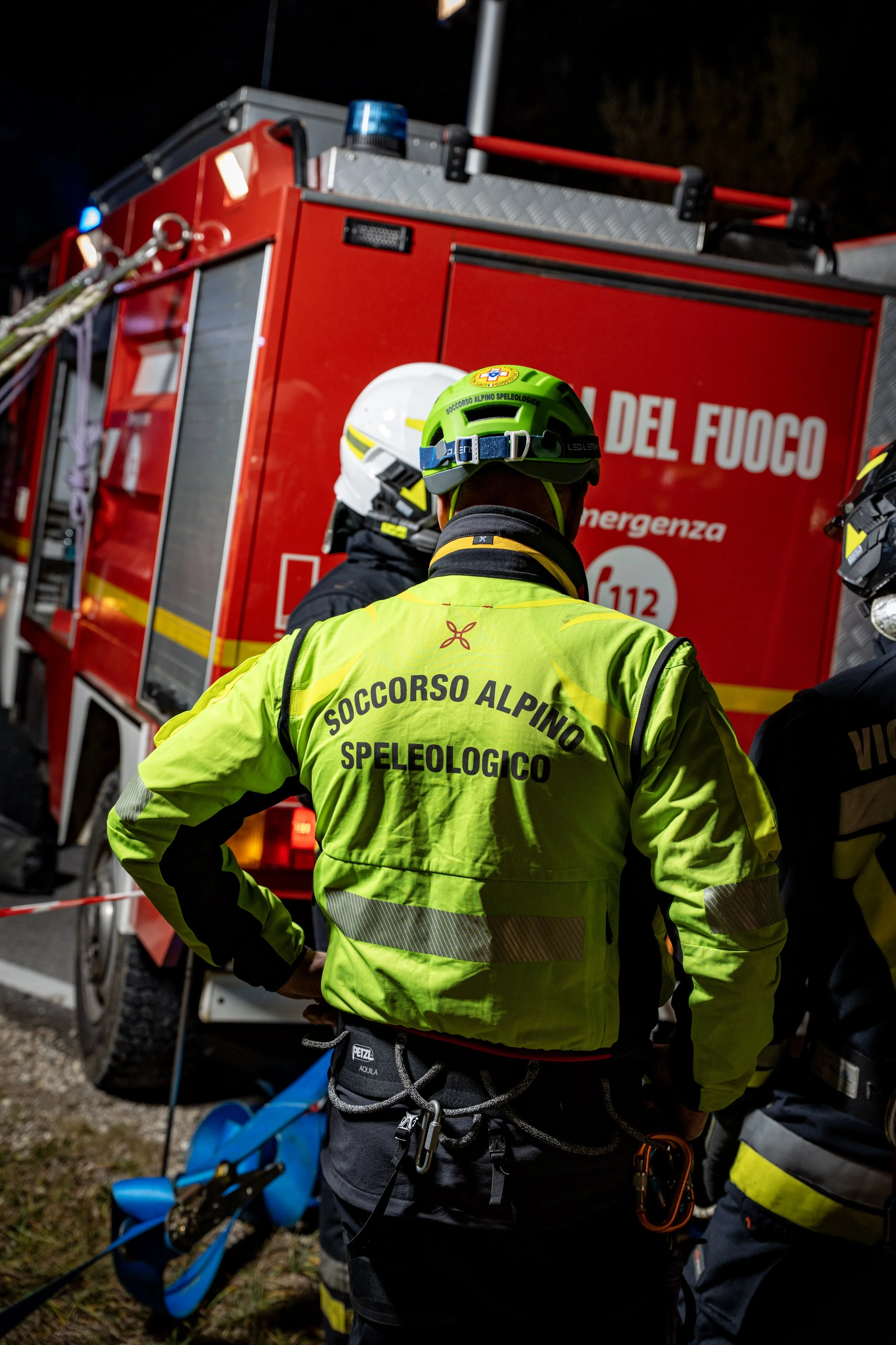 Emergency responders from Soccorso Alpino Speleologico wearing high-visibility jackets and helmets near a red fire truck at night.