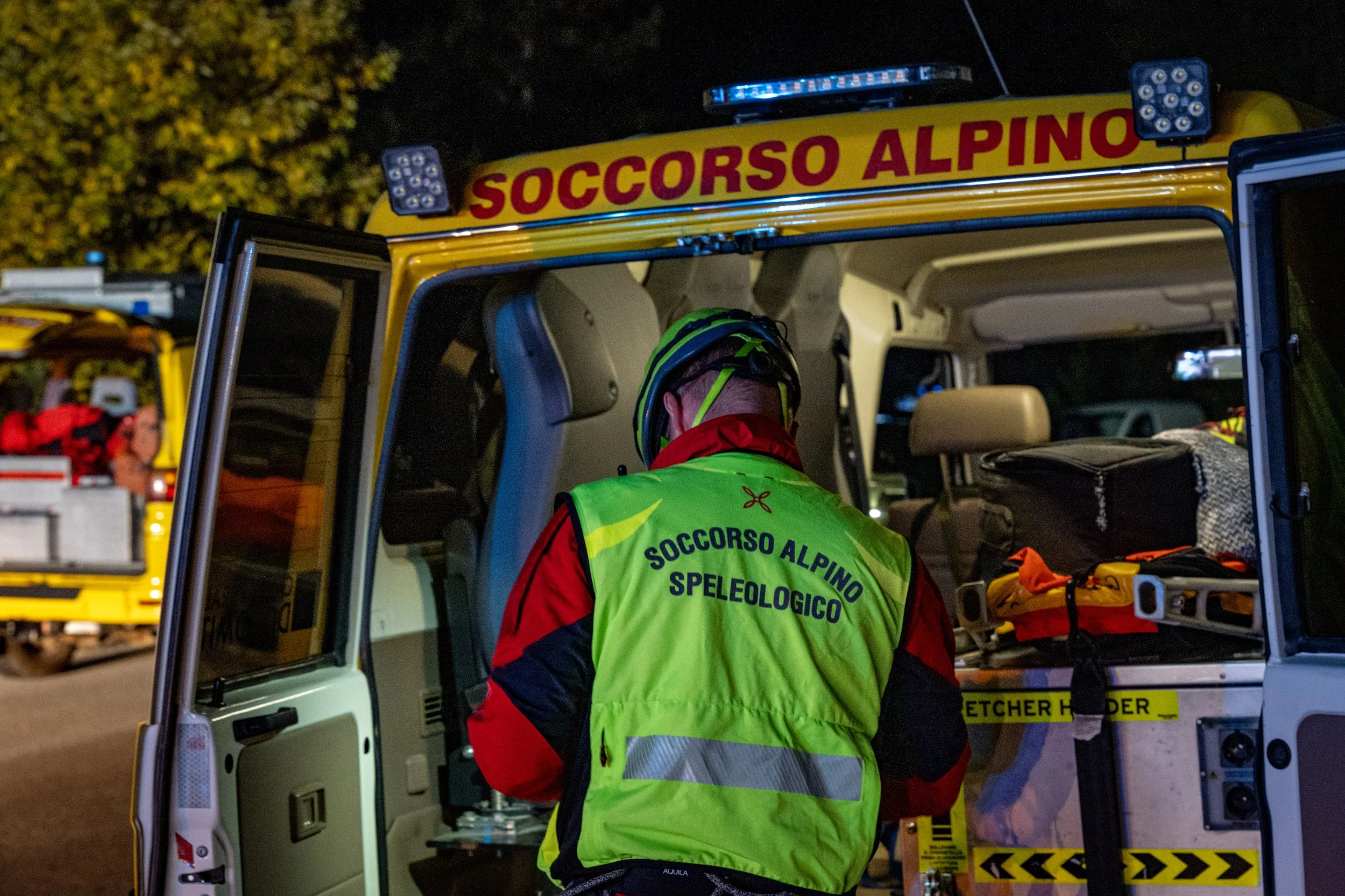 Rescue worker wearing a green vest with 'SOCCORSO ALPINO SPELEOLOGICO' printed on the back, working at night next to a yellow emergency vehicle branded 'SOCCORSO ALPINO,' with equipment and supplies inside.