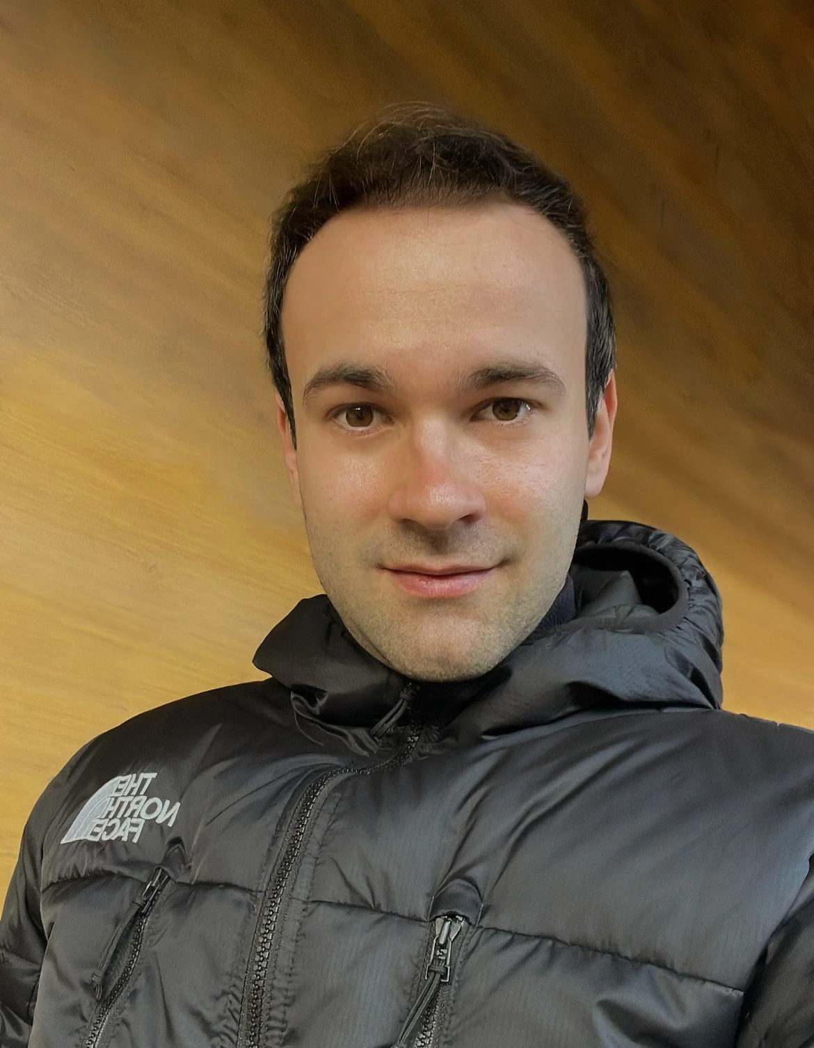 A man taking a selfie, wearing a black North Face jacket, with a wooden wall background.