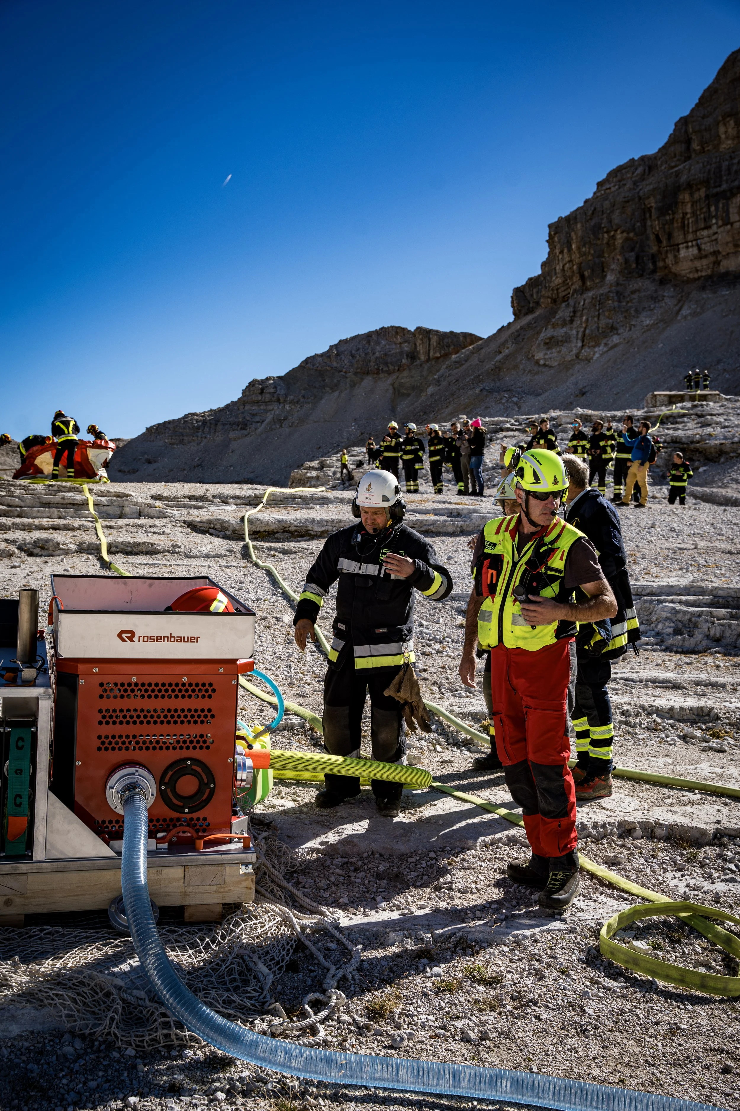 Firefighters and emergency personnel working on a rescue operation at a rocky, mountainous site under clear blue sky, with equipment including hoses and machinery