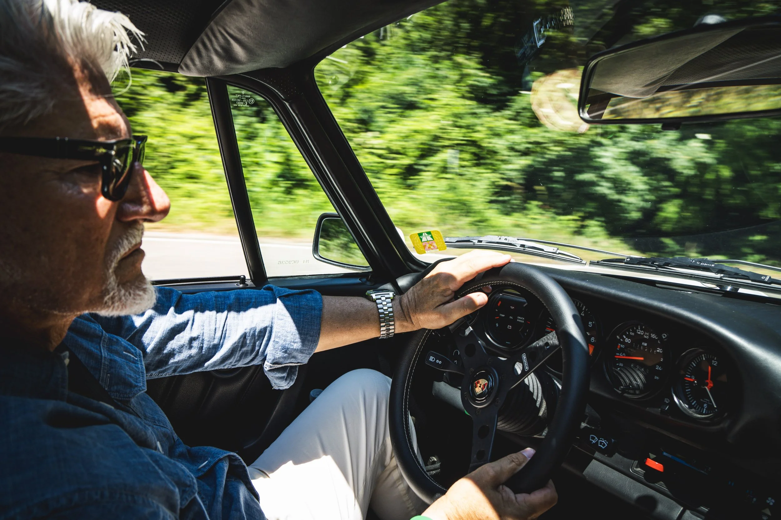 A man with white hair and sunglasses driving a black Porsche on a road surrounded by lush green trees.