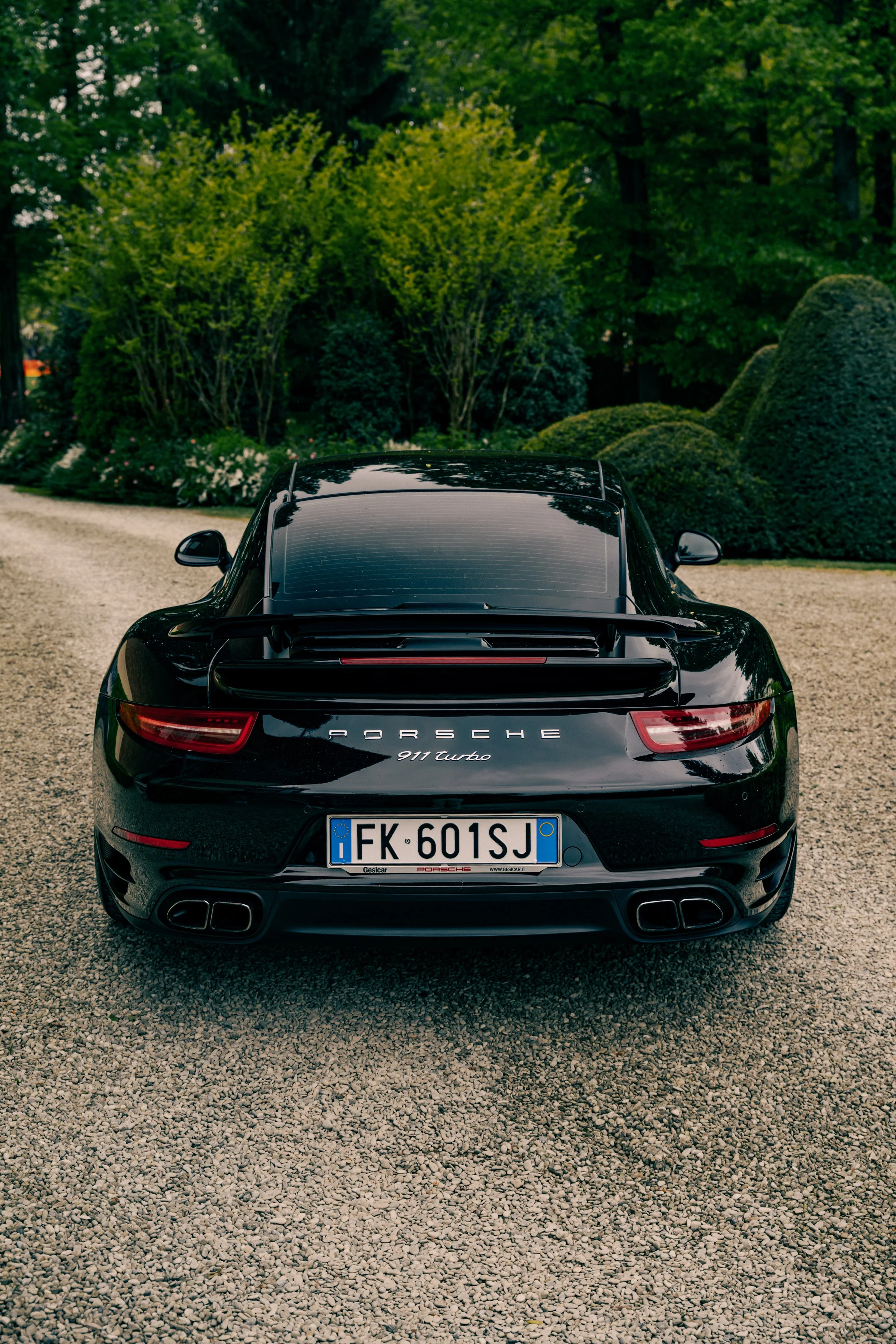 Black Porsche 911 Turbo parked on a gravel driveway with lush green trees and bushes in the background.