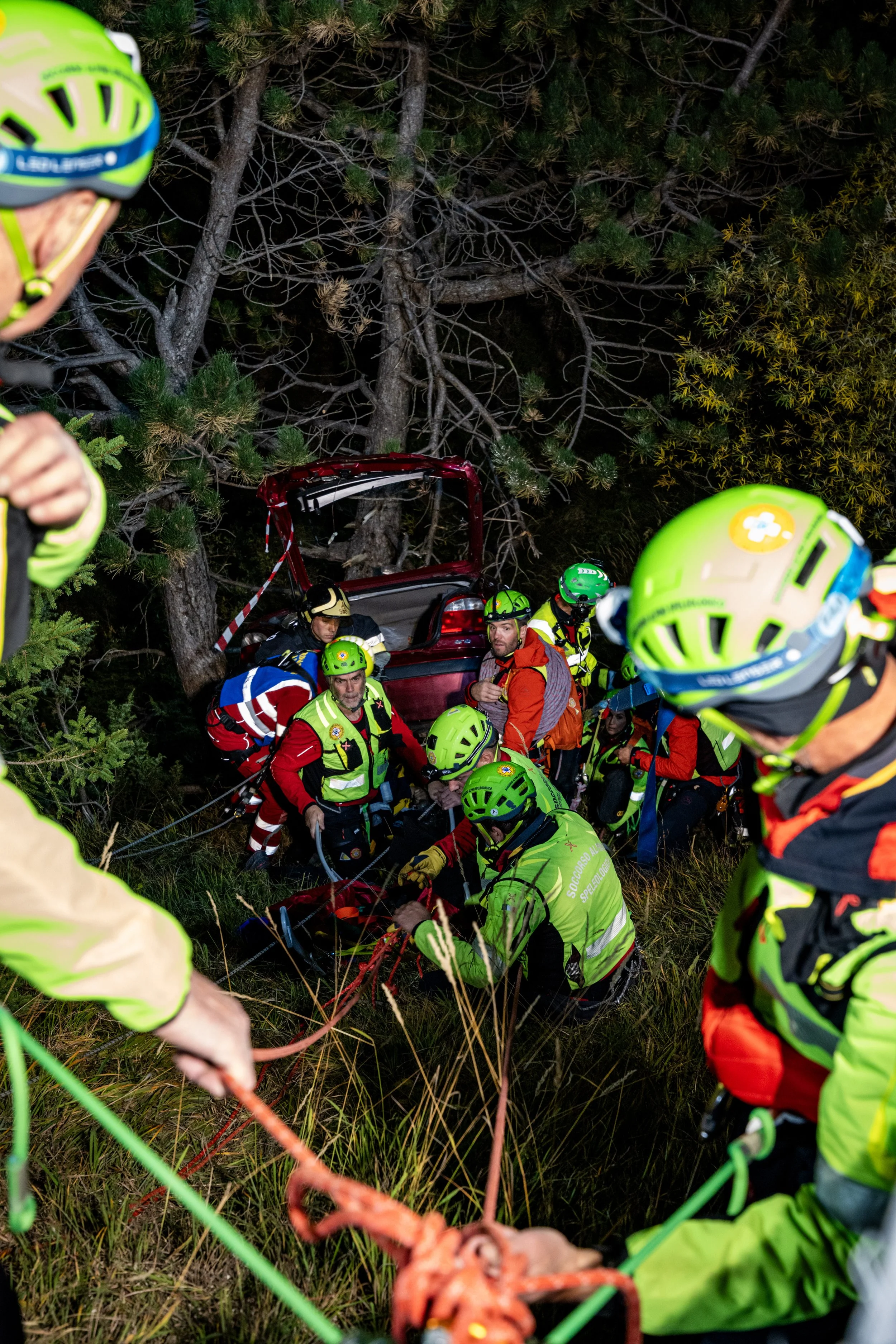 Nighttime rescue scene with multiple responders in high-visibility gear working around a downed vehicle in a forested area, with a tree and foliage in the background.