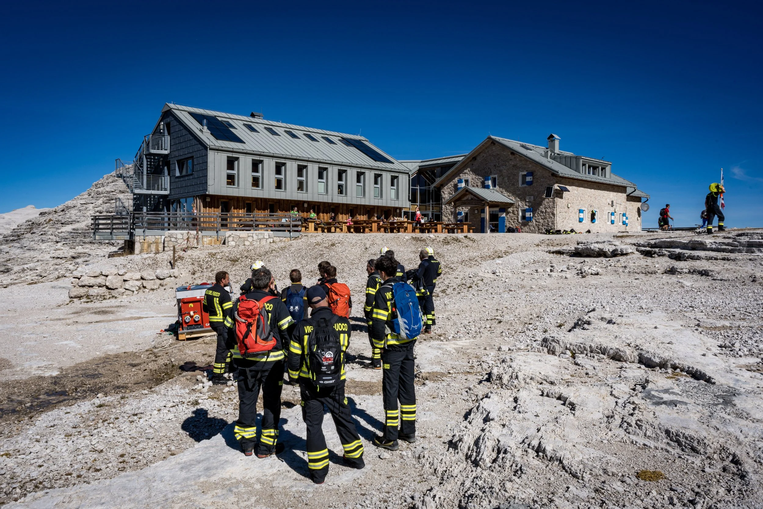 Group of rescue workers in black uniforms with yellow reflective stripes on rocky terrain outside a mountain cabin under a clear blue sky.