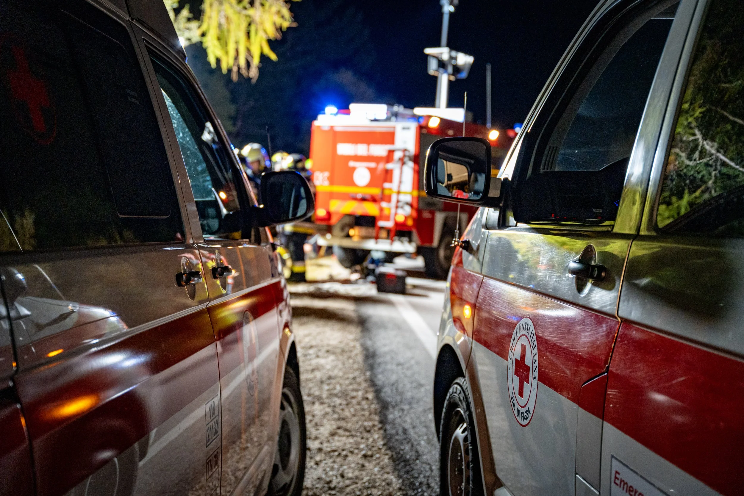 Nighttime scene of emergency responders attending to an incident between two parked cars, with a fire truck and emergency personnel visible in the background.
