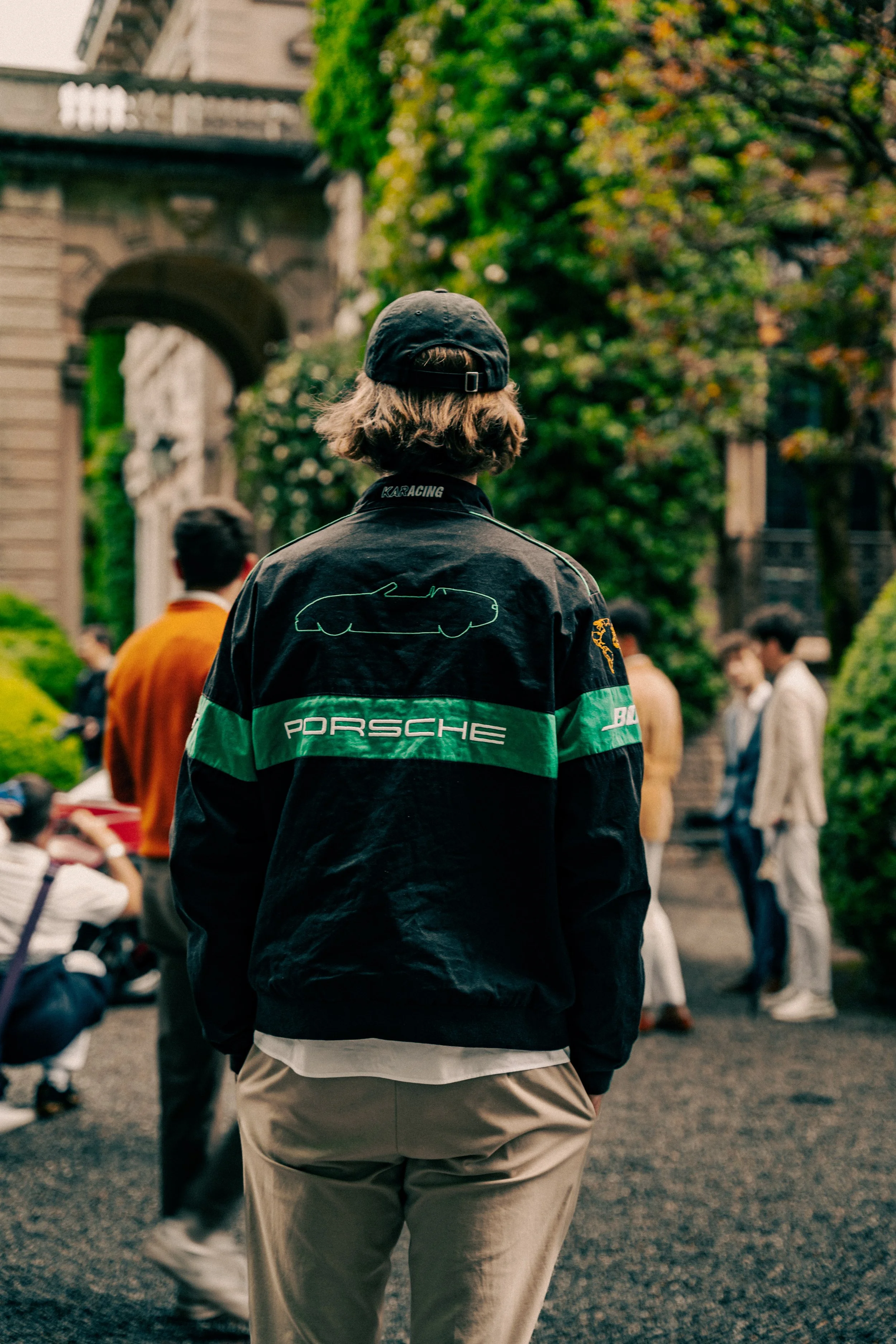 Person with long hair wearing a black cap and a black Porsche racing jacket standing outdoors, with a group of people and a building with arched doorways and greenery in the background.