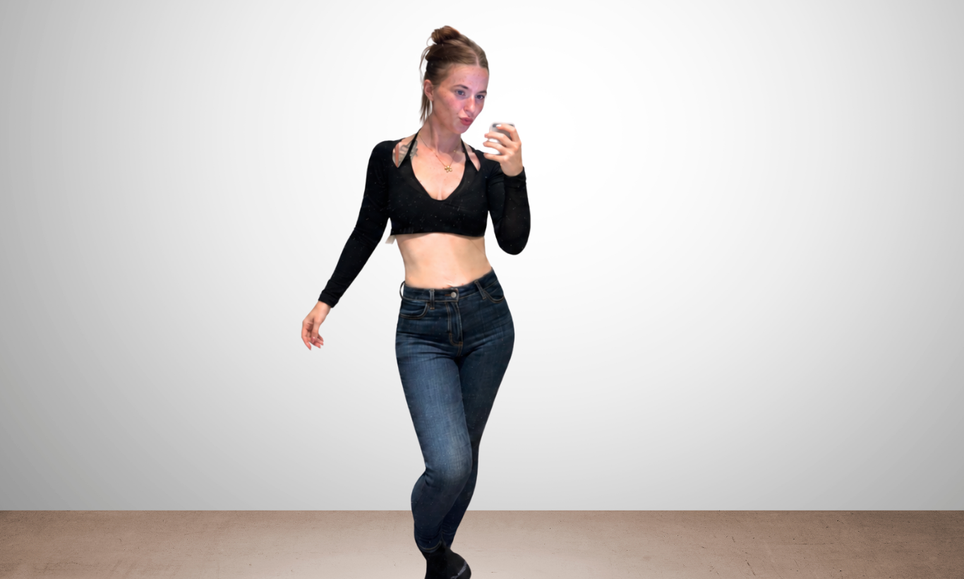 Woman taking a mirror selfie, wearing a black long-sleeve crop top and dark blue jeans, standing in front of a plain light gray wall.