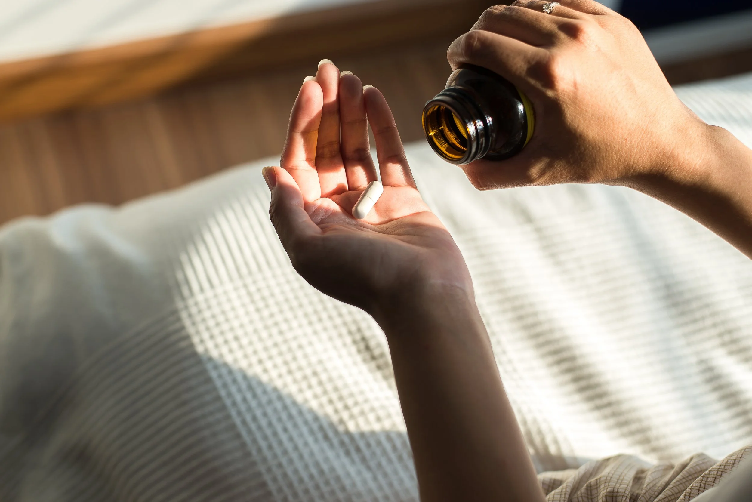A person holding an open brown glass bottle pouring a white capsule pill into their palm, with a soft-focus background.