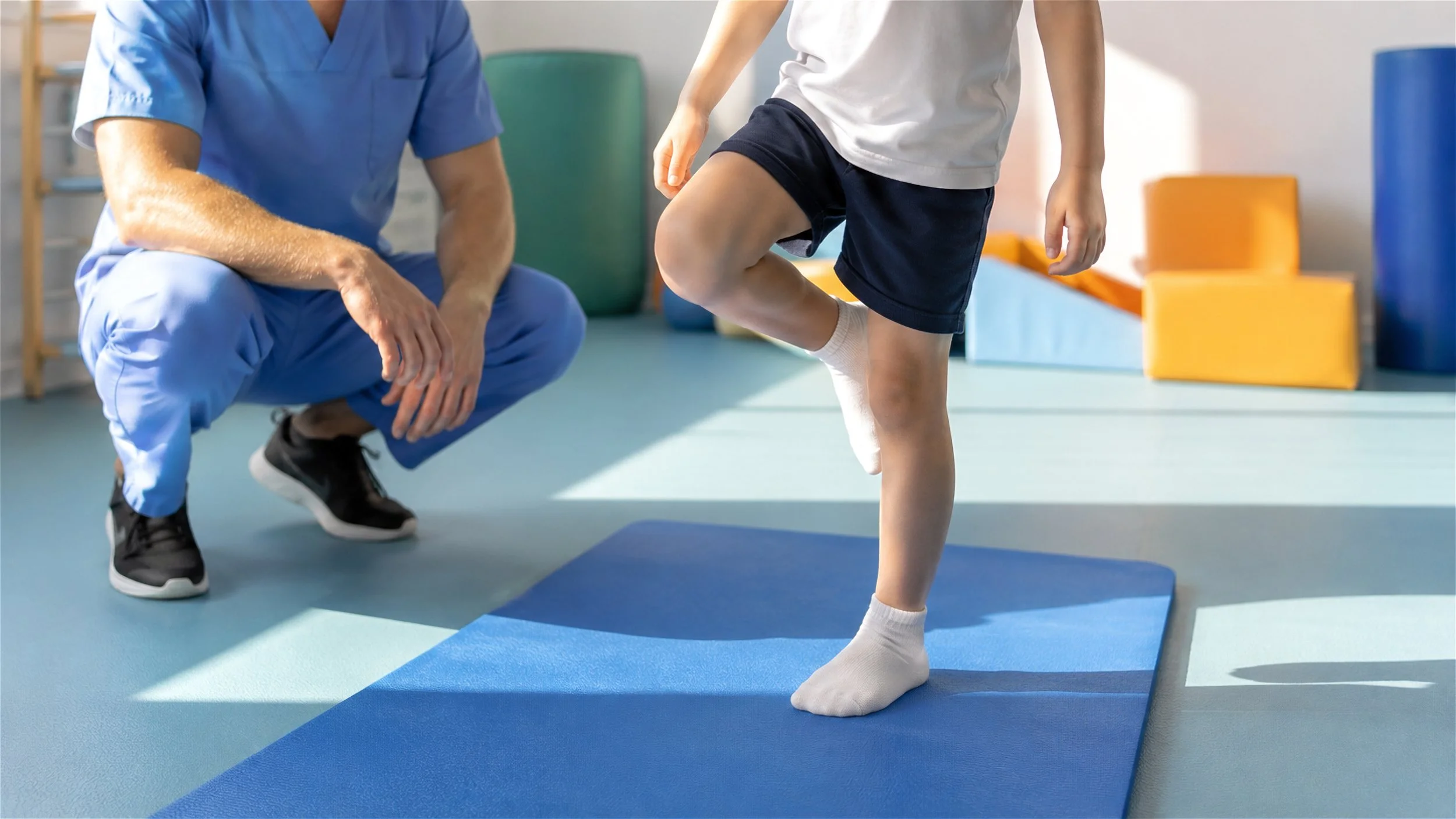 A child practicing balance on a blue yoga mat with a physical therapist or occupational therapist watching, in a therapy gym with colorful mats and equipment.