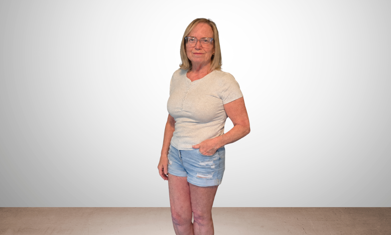 An older woman with blonde hair, wearing glasses, a light gray t-shirt, and ripped denim shorts, standing with one hand in her pocket against a plain white wall background.
