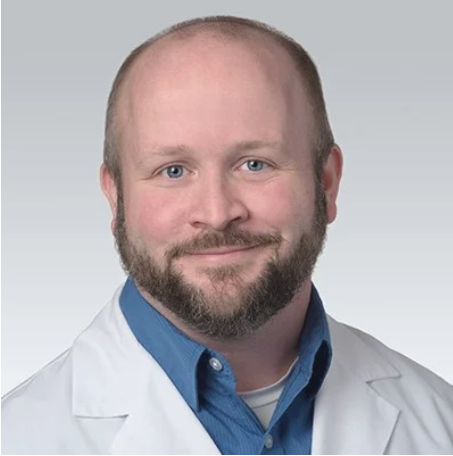 A man with a beard and short hair wearing a blue shirt and white lab coat, smiling at the camera.