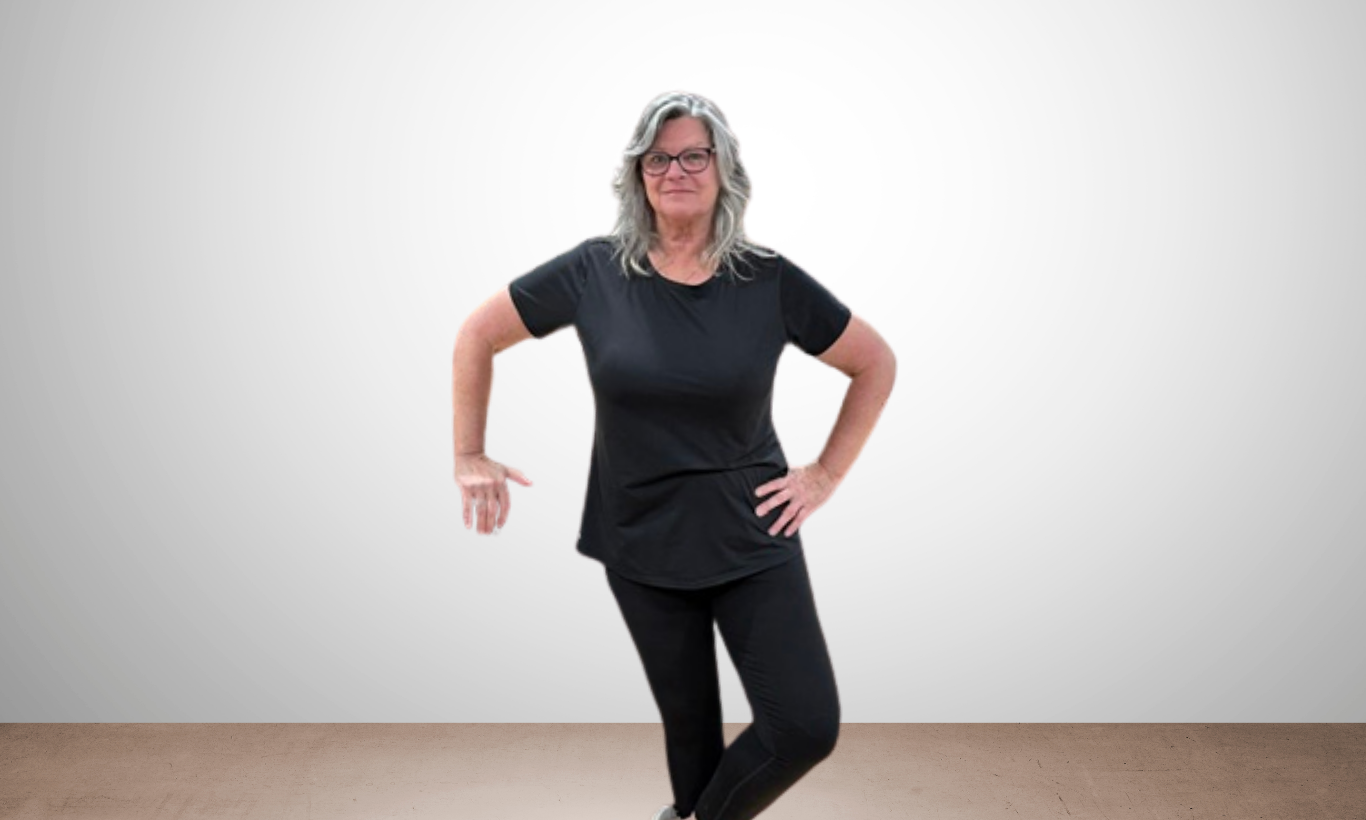 A middle-aged woman with gray hair and glasses standing indoors against a plain gray wall, wearing a black t-shirt and black pants, with one hand on her hip and the other extended downward.