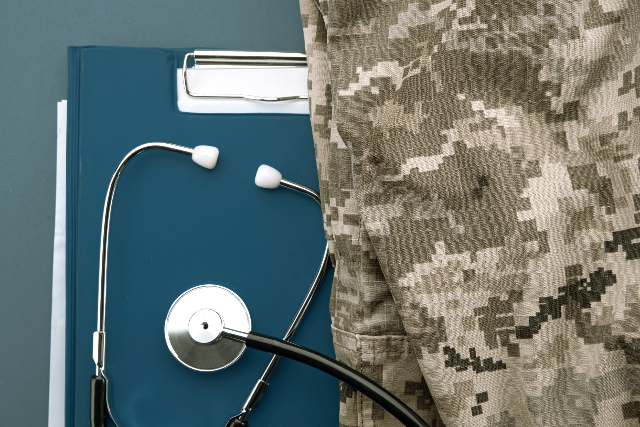 Close-up of a stethoscope on a blue folder next to a person wearing camouflage military uniform.