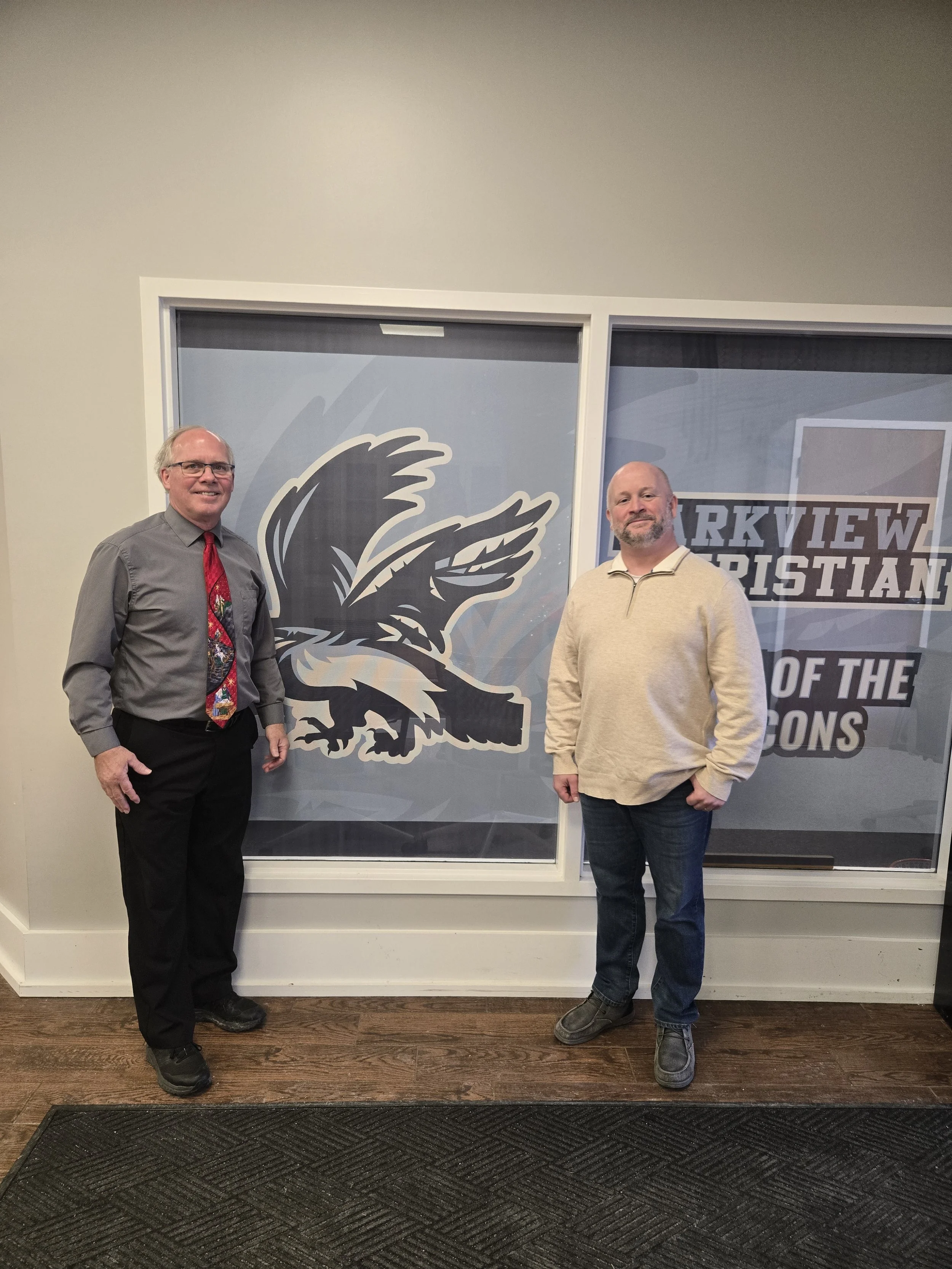 Two men standing in front of a window with a logo and text, wearing casual clothing, indoors with wooden floor and gray wall.