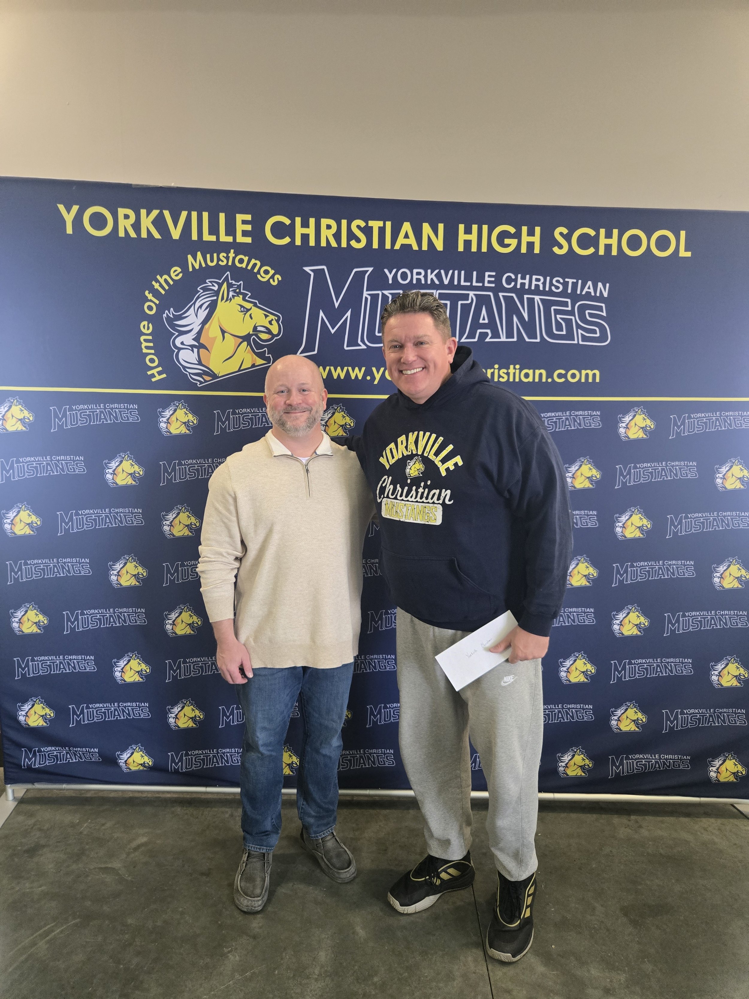 Two men standing together in front of a Yorkville Christian Mustangs backdrop, smiling at the camera.
