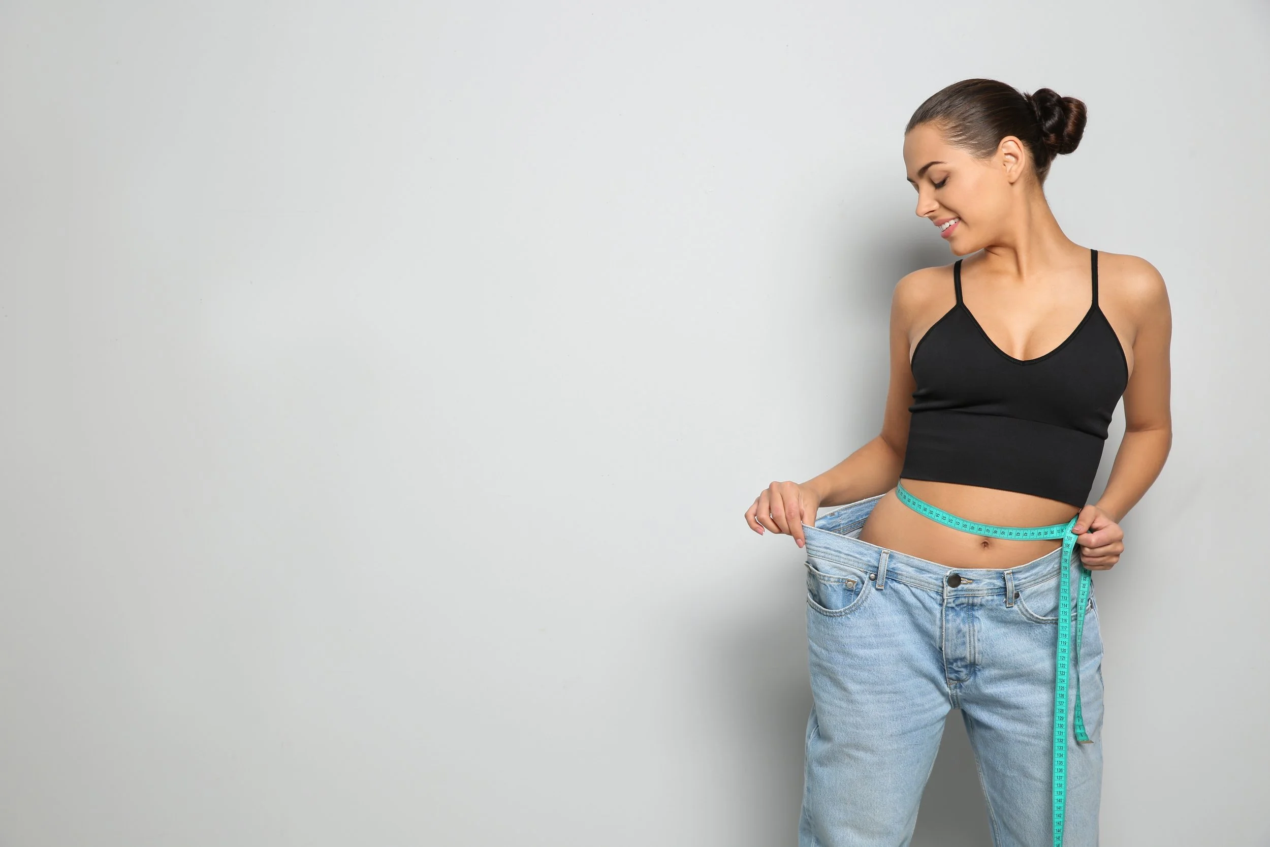 A woman standing against a plain gray background, wearing a black sports bra and loose blue jeans, measuring her waist with a blue measuring tape, and smiling while looking down.