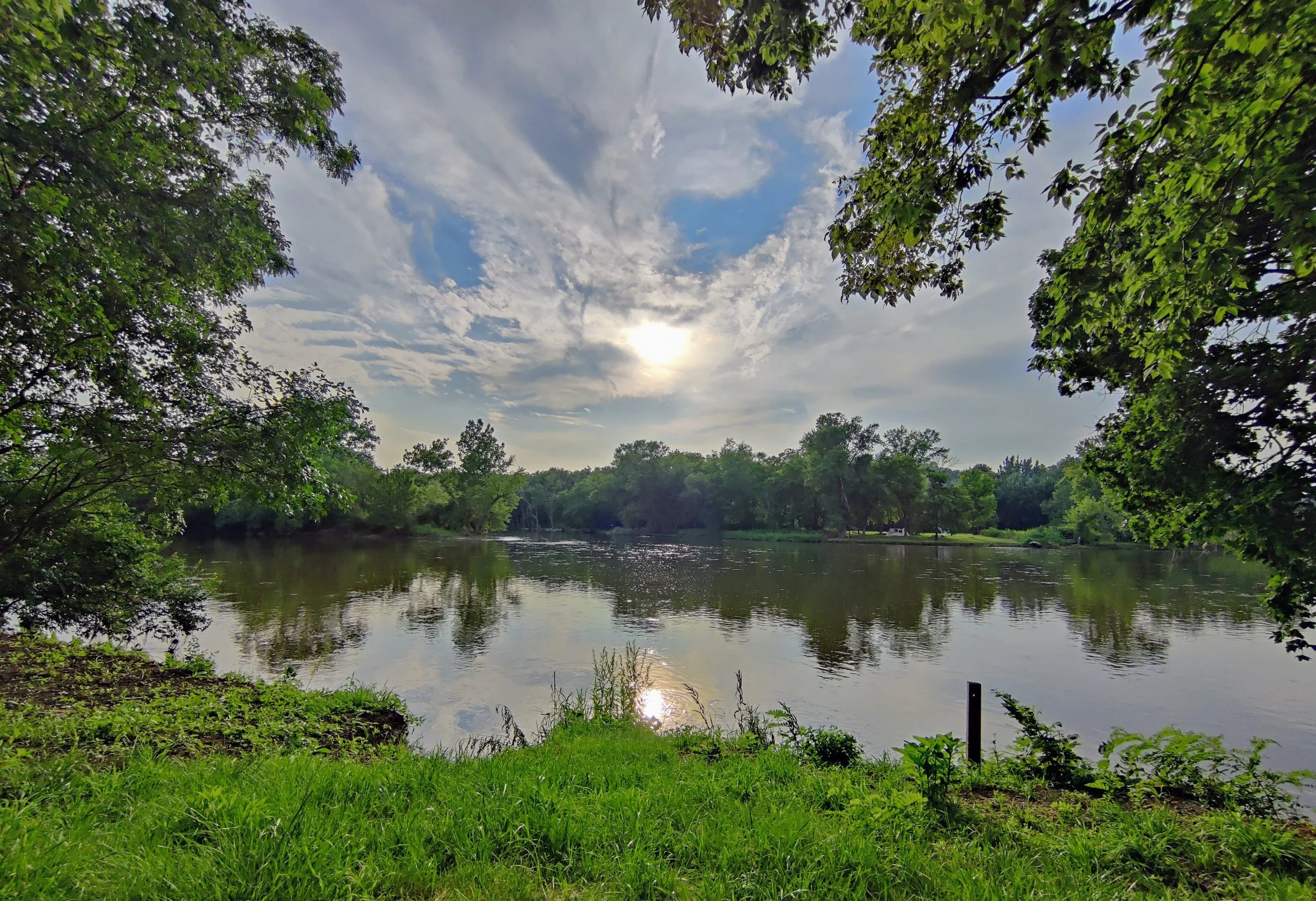 A peaceful scene of a river surrounded by green trees under a partly cloudy sky with the sun visible through the clouds.