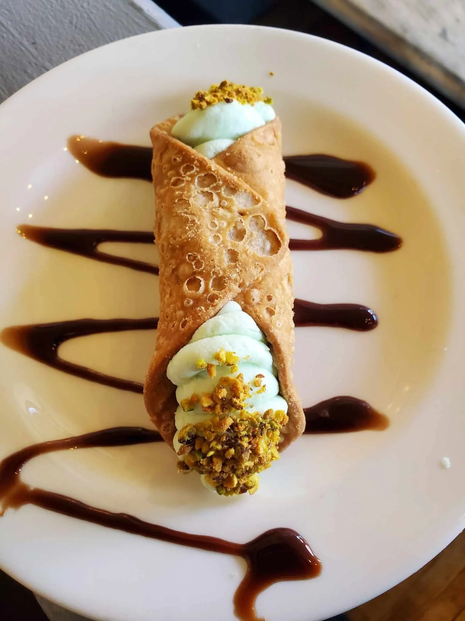 An ice cream cone on a white plate decorated with chocolate sauce, topped with crushed pistachios and a yellow garnish.