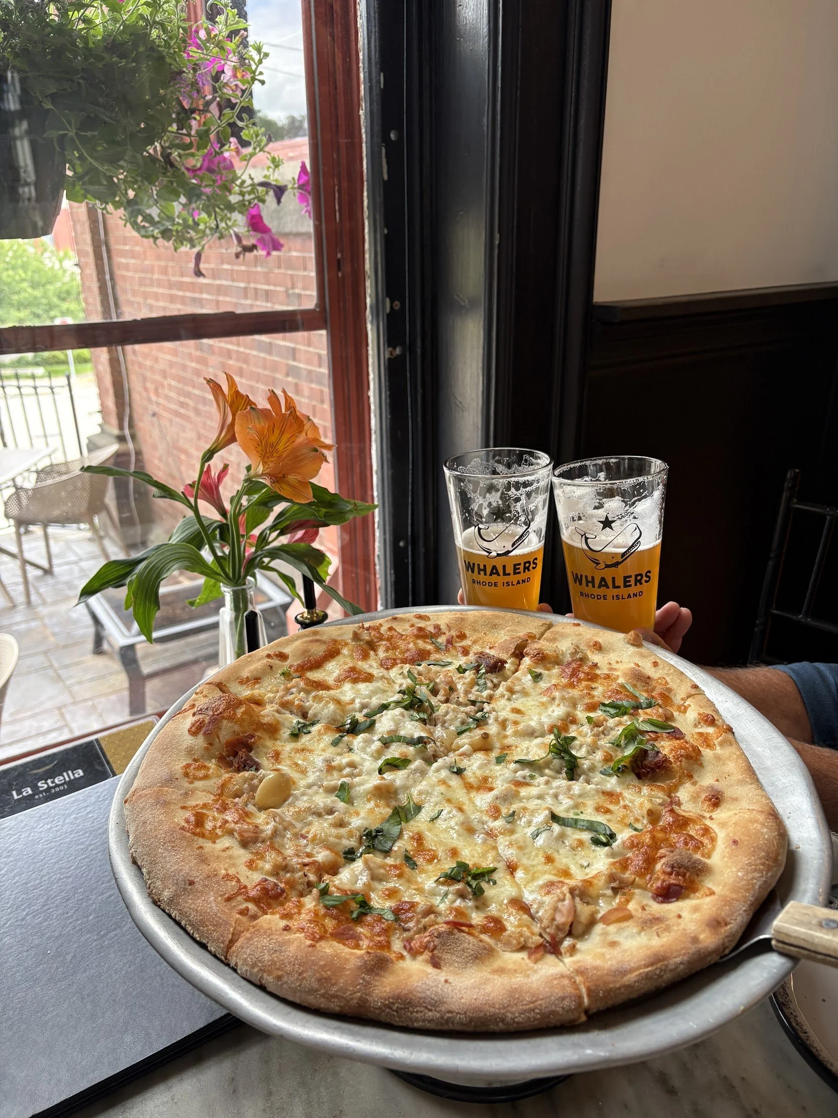 A large cheese pizza with green herbs on top, resting on a pizza tray. In the background, there is a window with pink and purple flowers and a brick wall outside. Two glasses of beer with