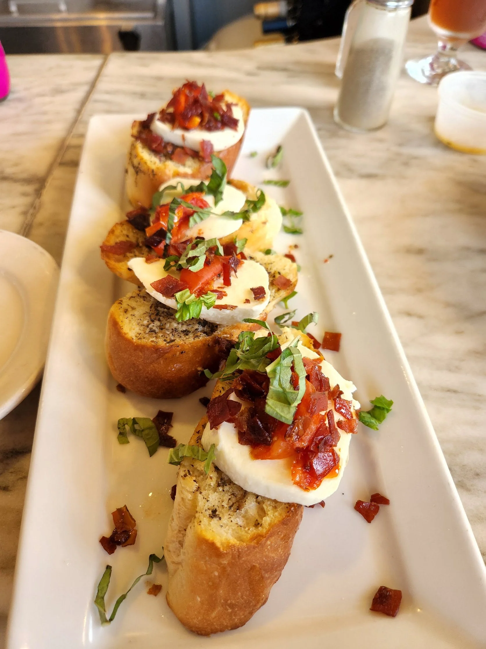 Three slices of bread topped with mozzarella cheese, tomato sauce, chopped bacon, and chopped fresh basil, served on a rectangular white plate.