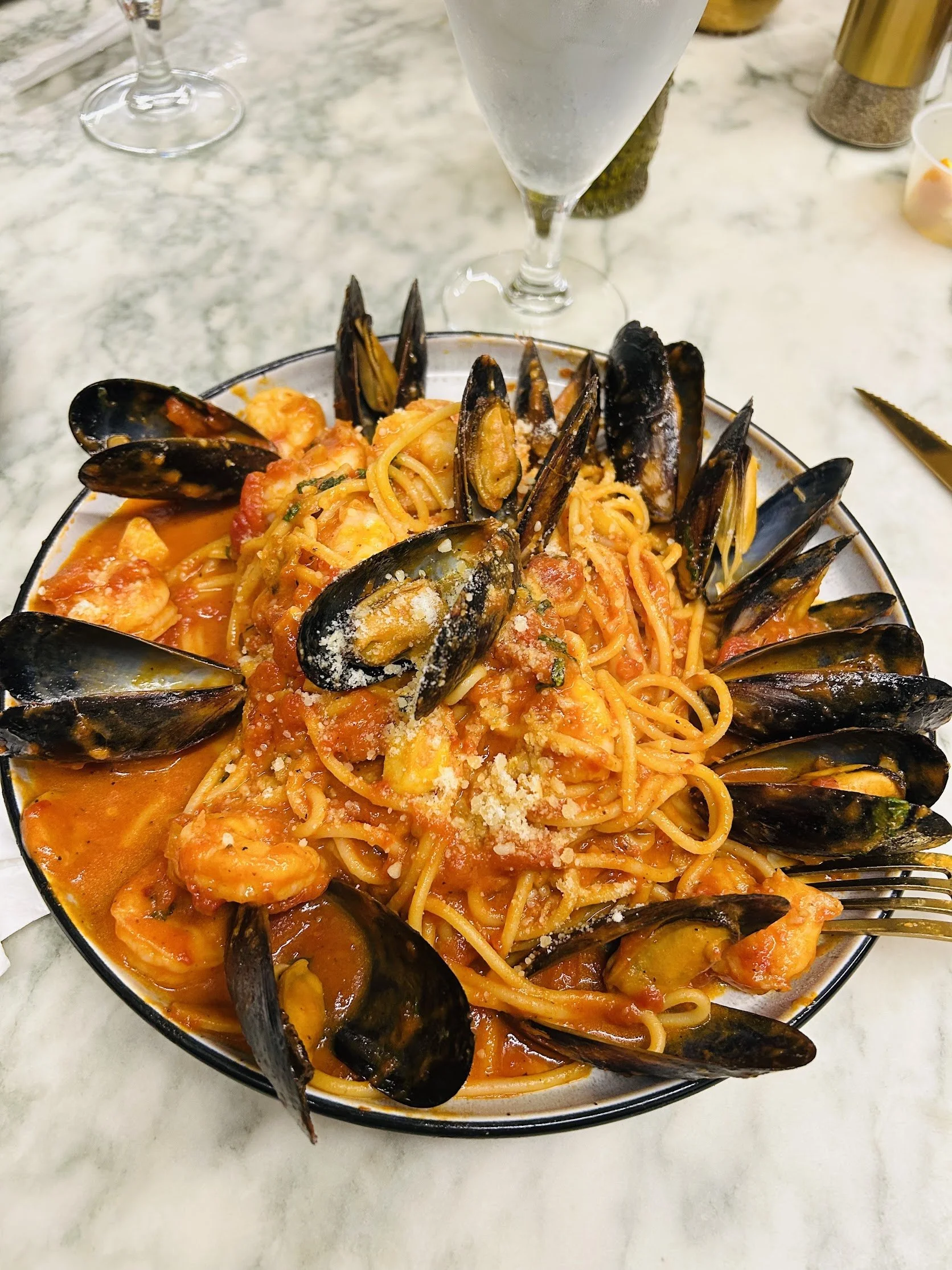 A bowl of seafood pasta with mussels, shrimp, and tomato sauce, topped with grated cheese, served on a marble table with a glass of water and condiments in the background.
