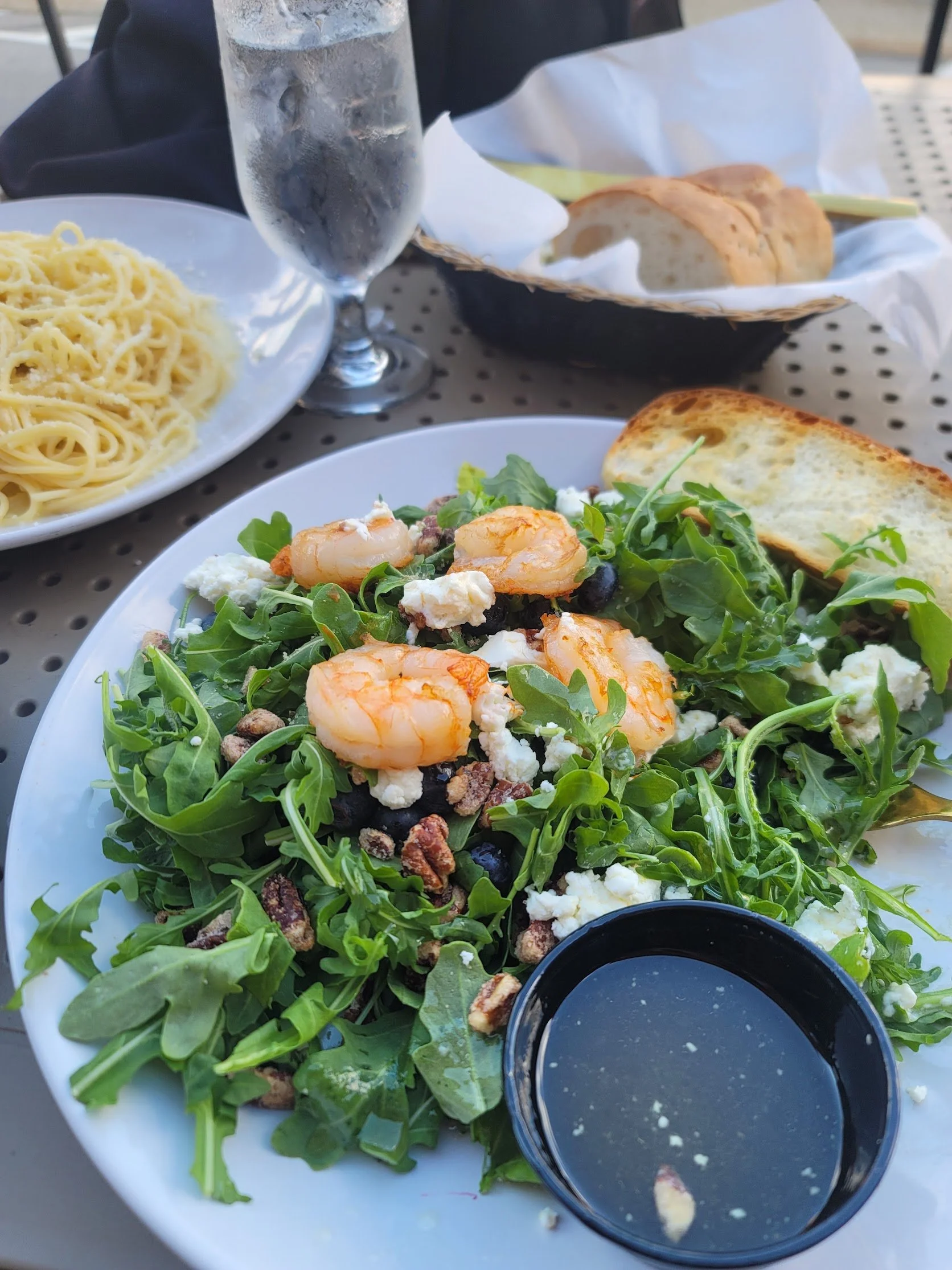 Close-up of a fresh green arugula salad with shrimp, feta cheese, and walnuts, served with a side of dressing. In the background, there is a plate of spaghetti, a glass of iced water, and a basket of sliced loaf bread on a table.