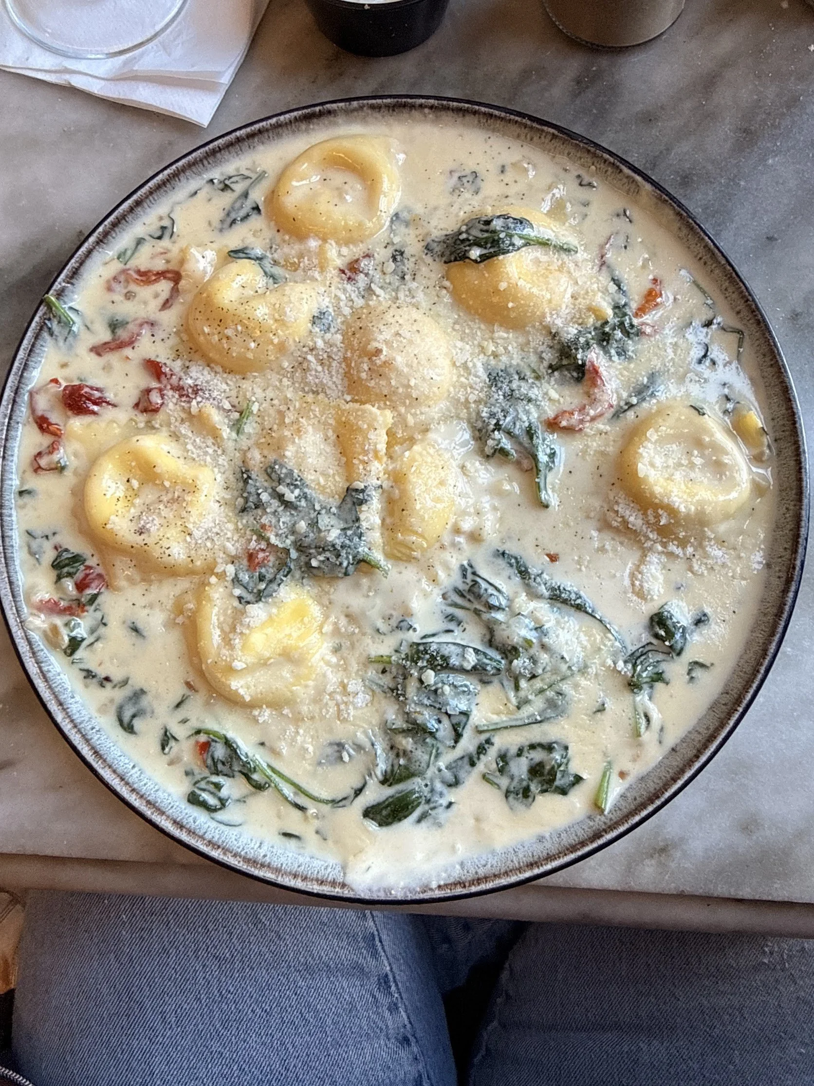 A bowl of creamy pasta with tortellini, spinach, and grated cheese on top. The bowl is placed on a table next to a glass of water, a black utensil holder, and a person wearing blue jeans.