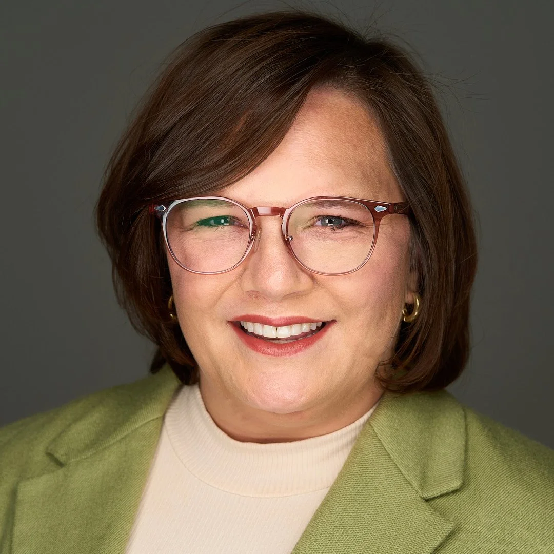 Close-up portrait of a smiling woman with short brown hair, wearing glasses, gold earrings, a yellow turtleneck, and a green blazer against a gray background.