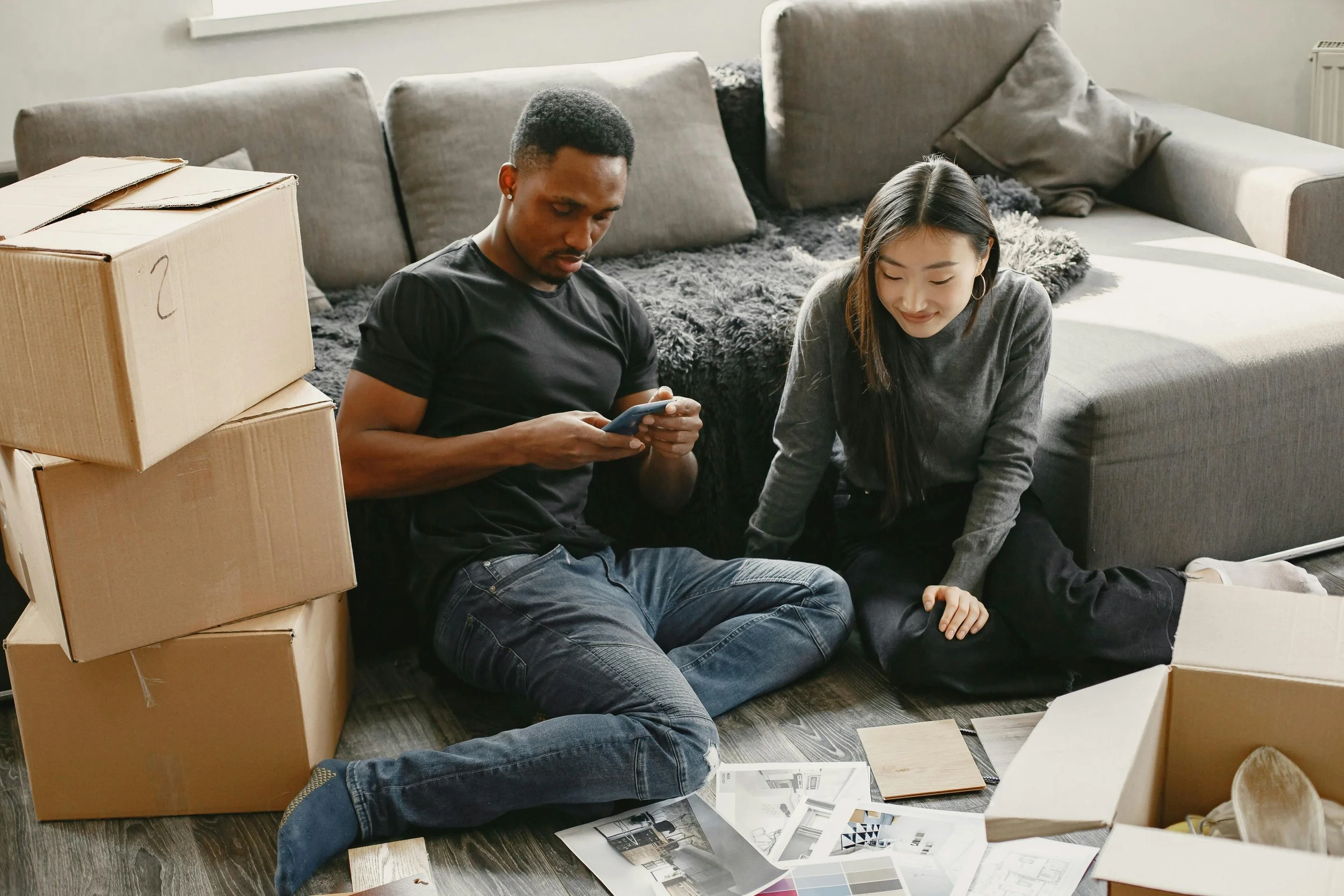 Two people sitting on the floor in a living room surrounded by cardboard boxes and interior design materials, with some papers, swatches, and a small open notebook spread out in front of them.