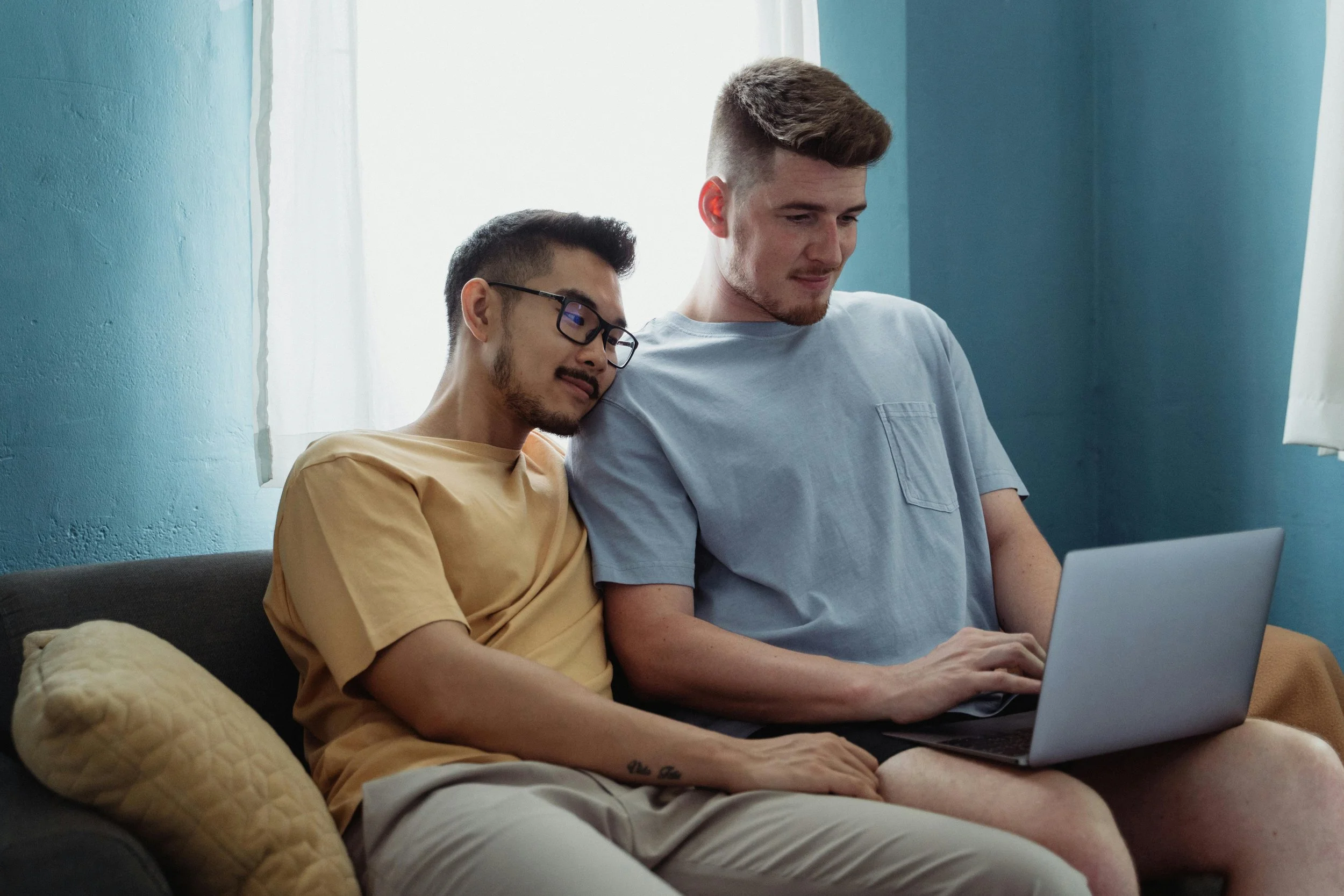 Two young men sitting on a couch, sharing a laptop together in a room with light coming through the window.
