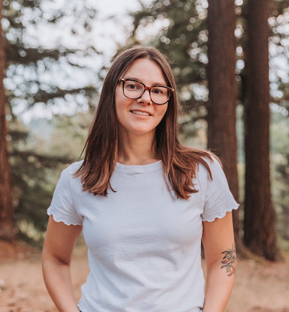 A young woman with brown hair, glasses, and a white t-shirt standing outdoors in front of tall trees.