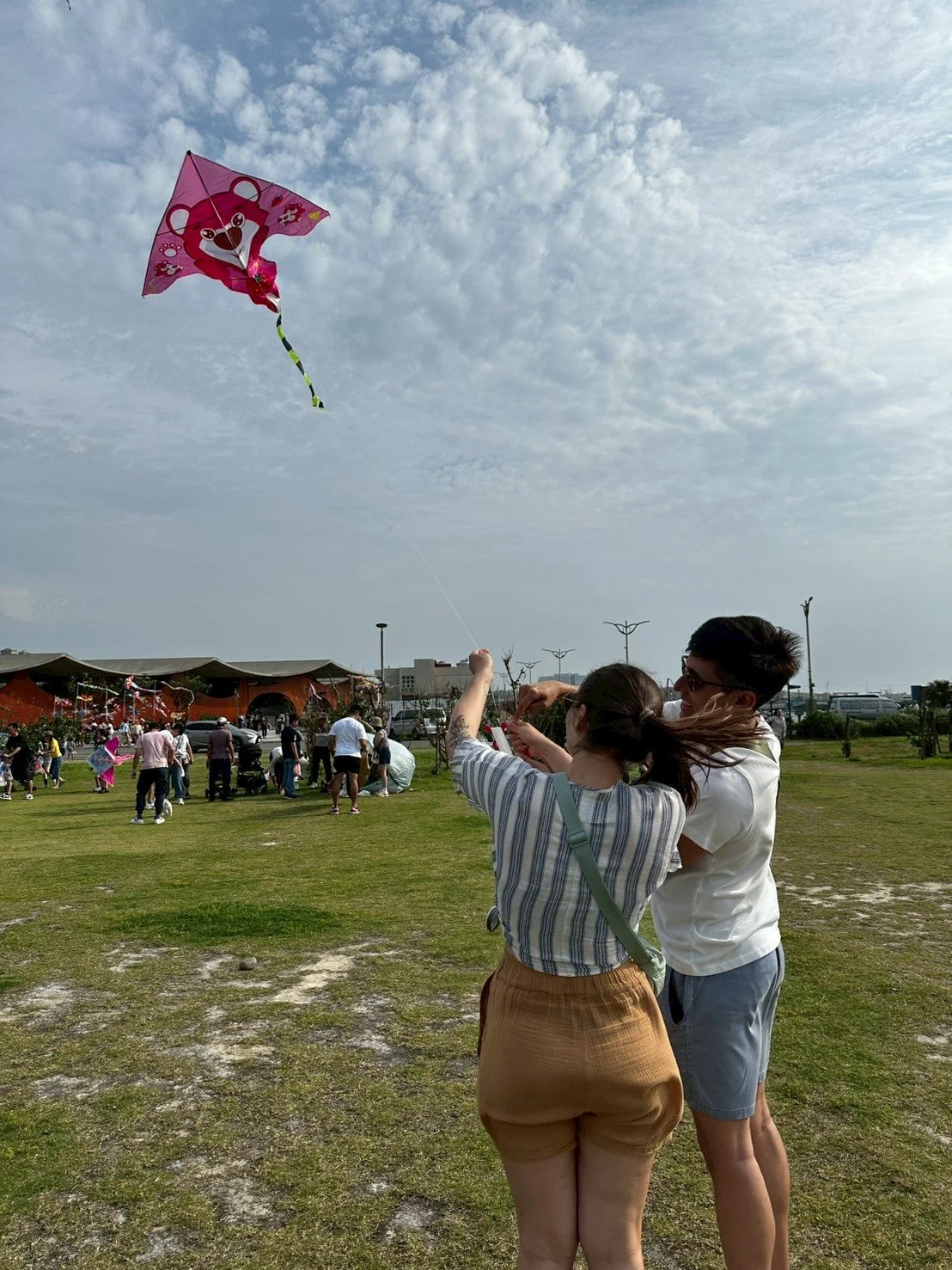 A group of people flying kites in a park on a cloudy day. Two people are in the foreground, one holding a pink bear-shaped kite while others are seen in the background enjoying the outdoor event.