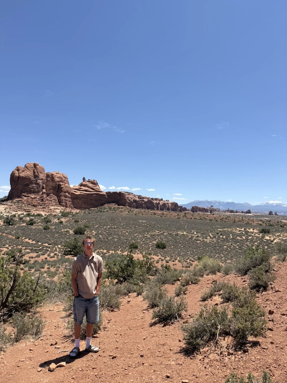 A young person standing on a dirt trail in a desert landscape with red rock formations, low shrubs, and mountains in the background under a clear blue sky.
