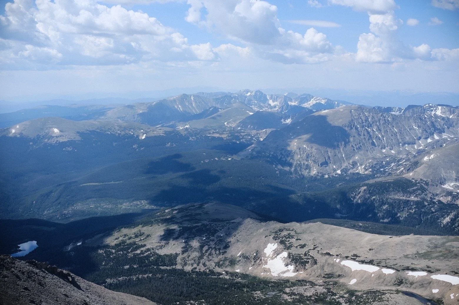 A vast mountain landscape showing rugged, snow-capped peaks under a partly cloudy sky with some cloud shadows on the slopes.
