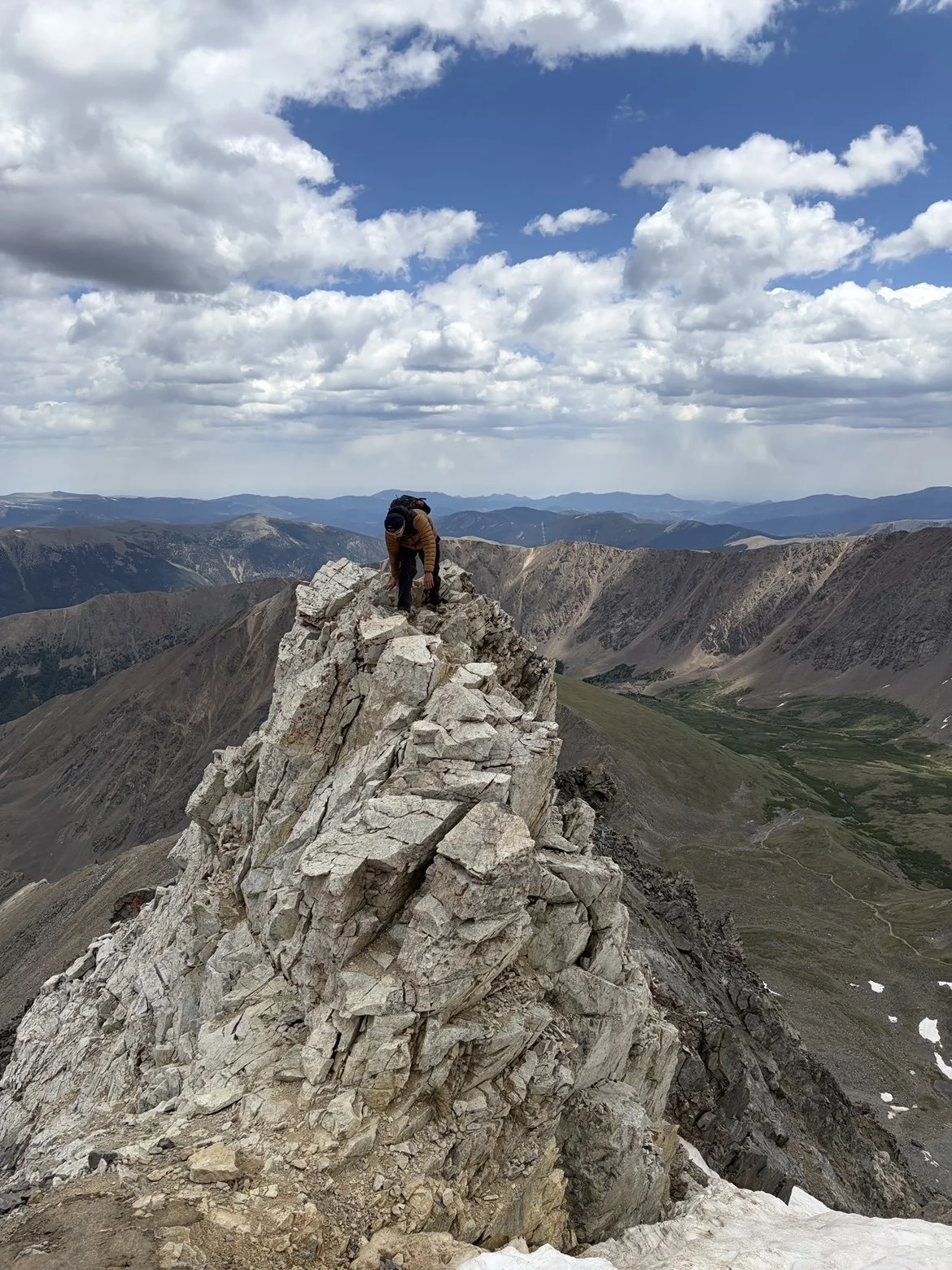 A person in outdoor gear standing on a jagged rocky mountain peak with expansive mountain ranges and a partly cloudy sky in the background.