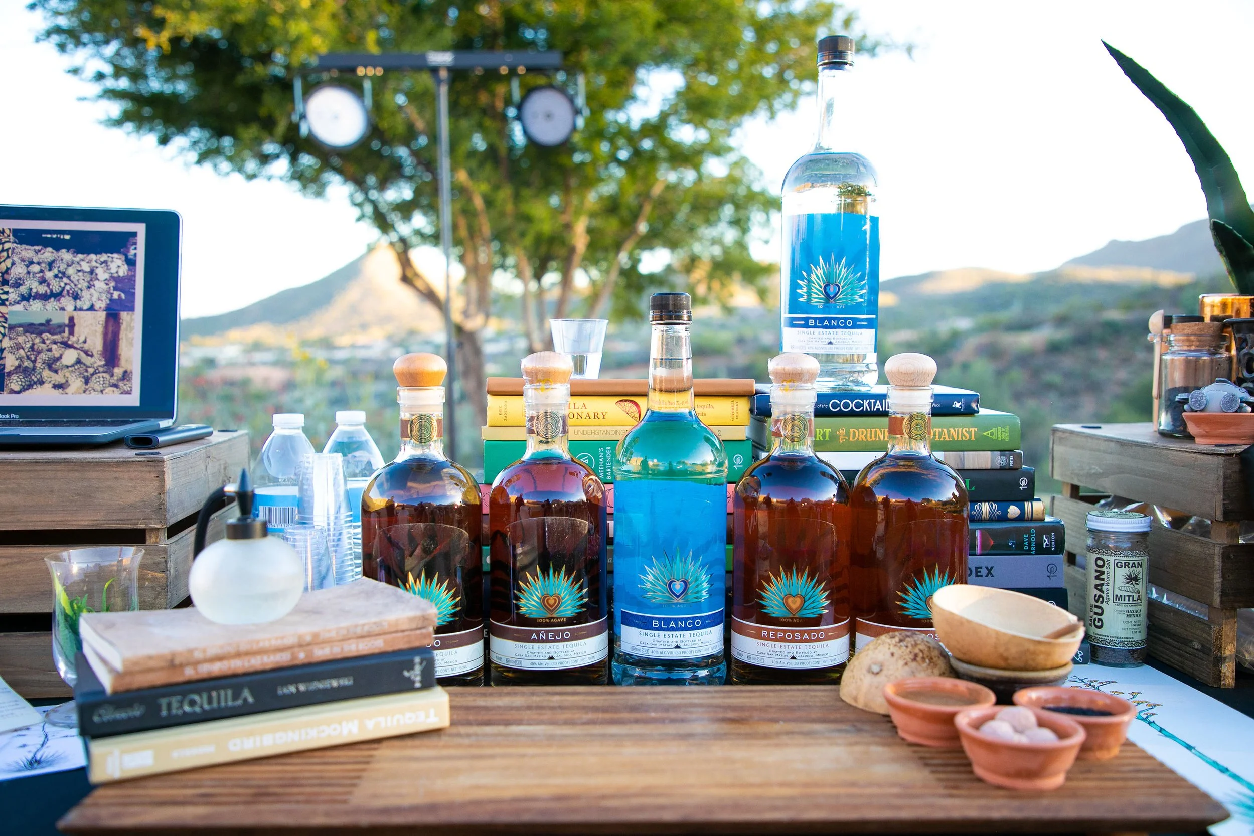 A table set outdoors with bottles of tequila, books, shot glasses, small clay dishes, and a laptop showcasing flower images, against a background of trees and mountains.