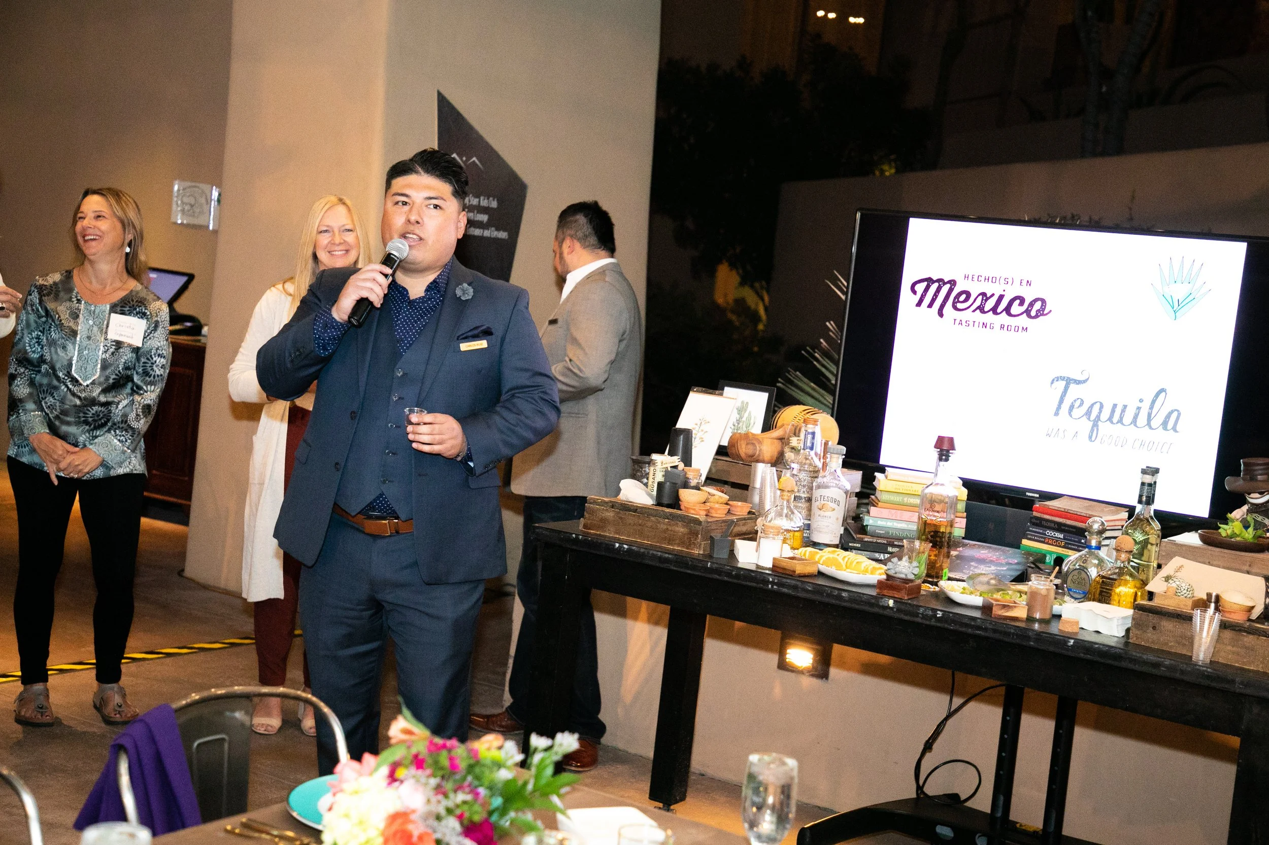 A man in a dark blue suit holding a microphone and a shot glass, speaking at a social event with women smiling in the background. There is a large screen displaying a logo for a tasting room in Mexico and various bottles and items on a table.
