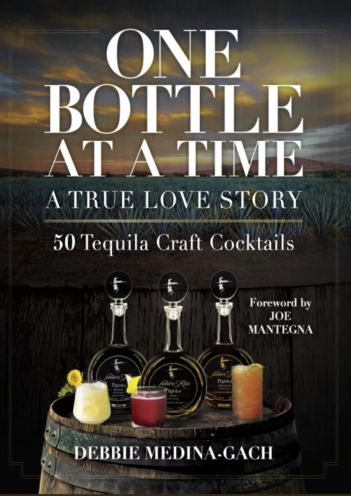 Book cover titled 'One Bottle at a Time: A True Love Story - 50 Tequila Craft Cocktails' with a sunset over a field in the background, featuring images of tequila bottles on a barrel and three colorful cocktails in front of them, authored by Debbie Medina-Gach with a foreword by Joe Mantegna.