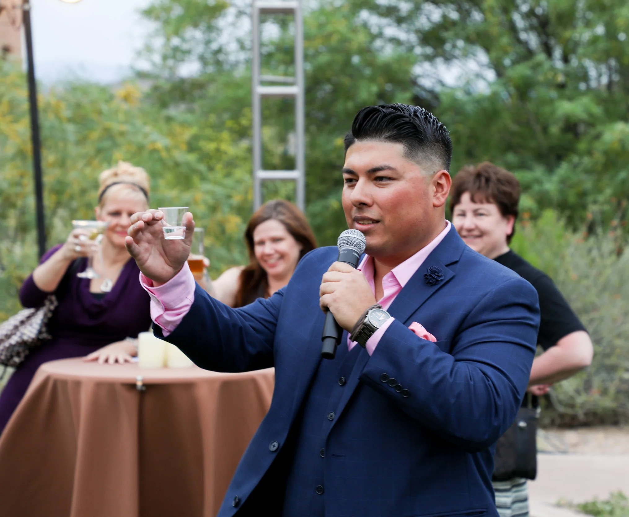 A man in a blue suit and pink shirt speaking into a microphone at an outdoor gathering, surrounded by people raising glasses for a toast, with trees and a metal scaffolding in the background.