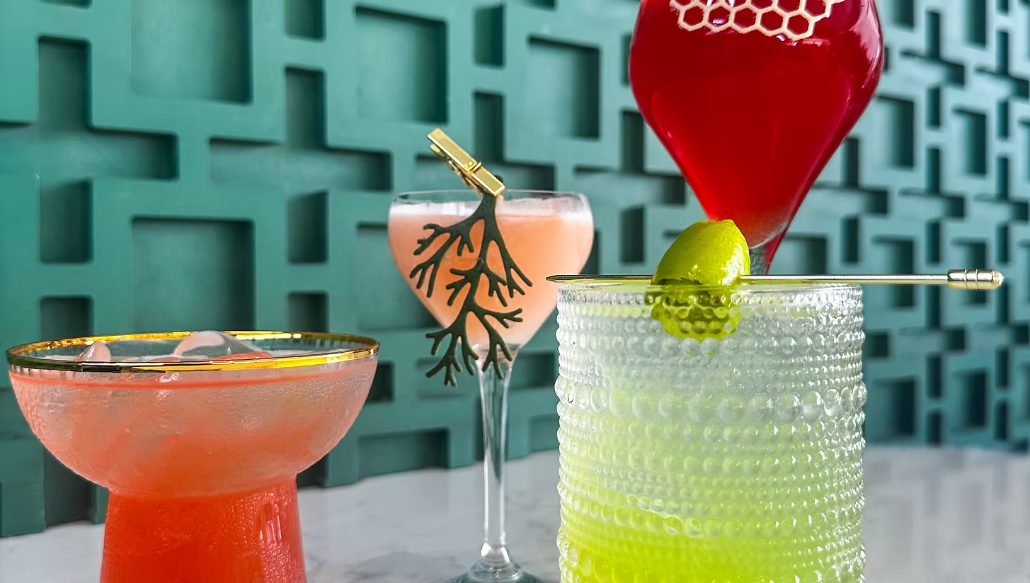 Four colorful cocktails on a table against a green geometric wall. One in a short glass with a red drink and ice, has a gold rim. Another in a tall spoon-shaped glass with a pink drink and black branch decoration. The third in a textured transparent glass with lime garnish and a cocktail pick, has a green drink. The last in a large red vase-shaped glass with a red drink.