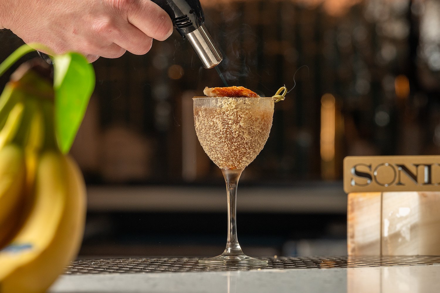 A person is pouring a flame torch over a cocktail in a clear wine glass, which is coated in a layer of sugar, with a slice of fruit on top.