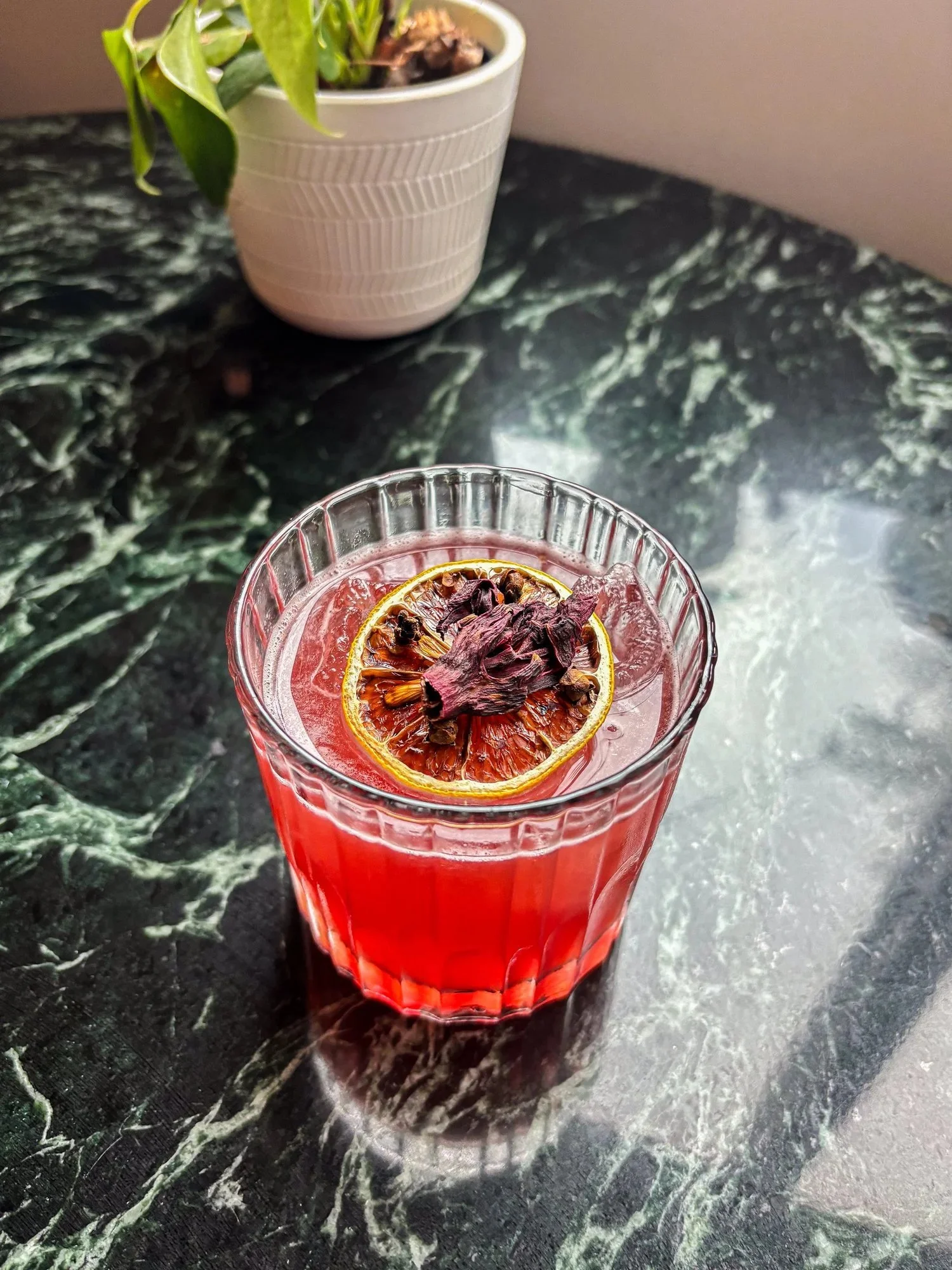 A red beverage in a tall, clear glass garnished with dried lemon slices and dried hibiscus flowers, placed on a dark green marble surface.
