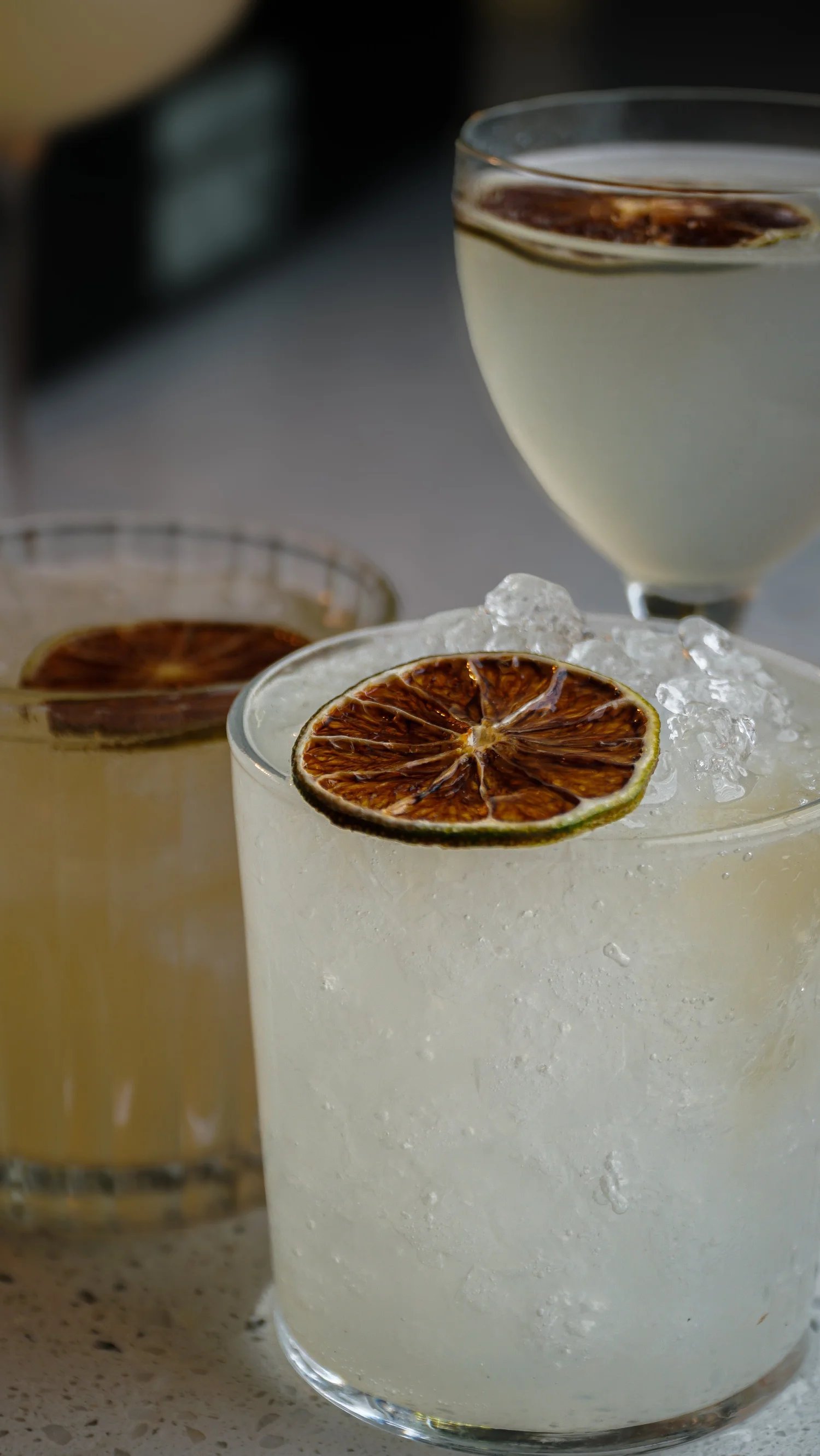 Three cocktails with dried citrus slices garnishing the rims, one with ice and a second with a dark drink in the background.