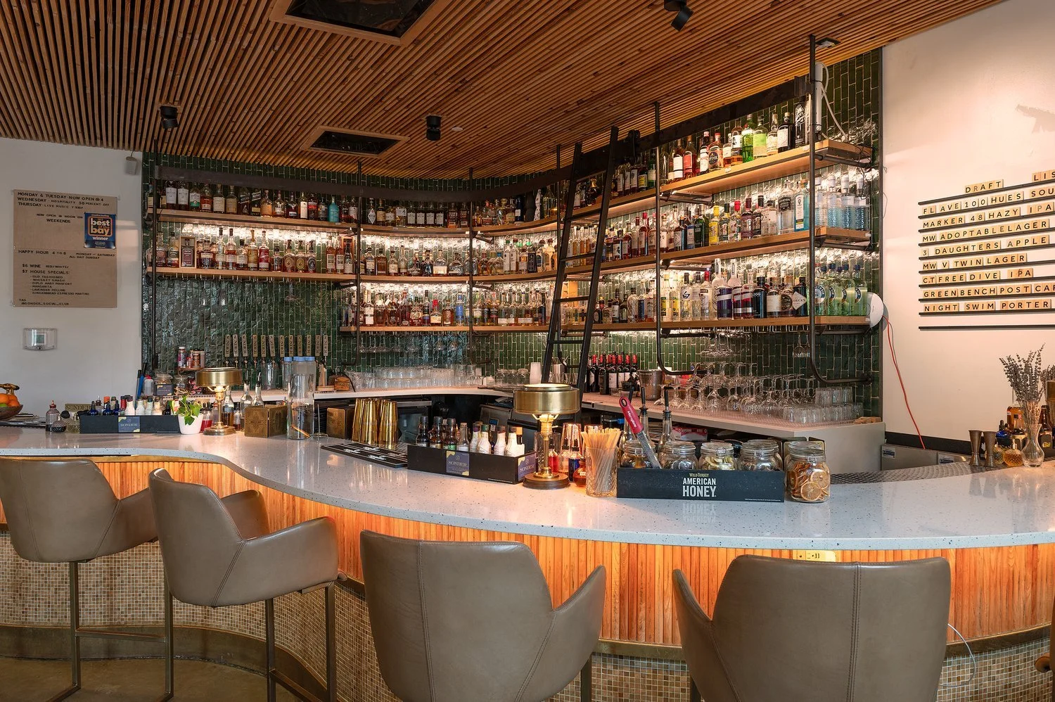 Inside a modern bar with a curved white countertop and beige chairs. Shelves filled with various bottles of liquor and spirits are behind the bar, with a green tile backsplash and a wooden ceiling.