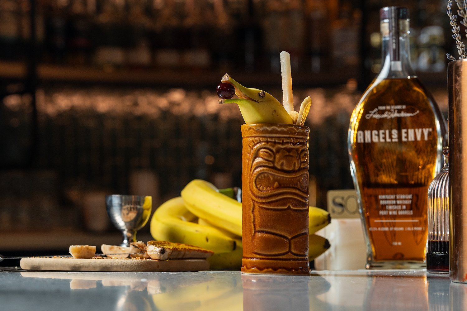 A tiki-style cocktail with banana, cherries, and fruit garnishes in front of a bunch of bananas, with a bottle labeled "Angels Envy" bourbon and a small stainless steel cup in the background.