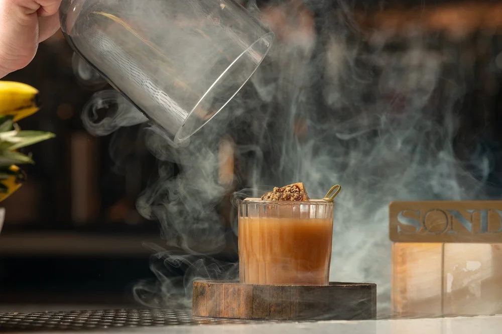 A glass of hot tea with steam rising, topped with a piece of cake and garnished with a small spoon, placed on a wooden coaster.
