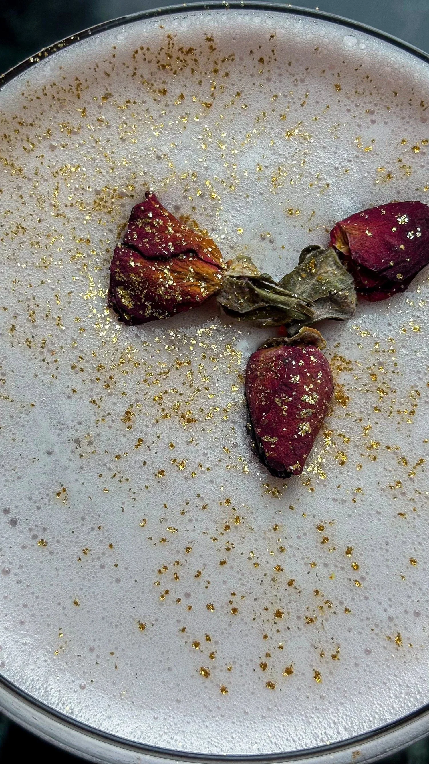 Three dried rosebuds and dried rose petals sprinkled with gold glitter on a foamy surface.