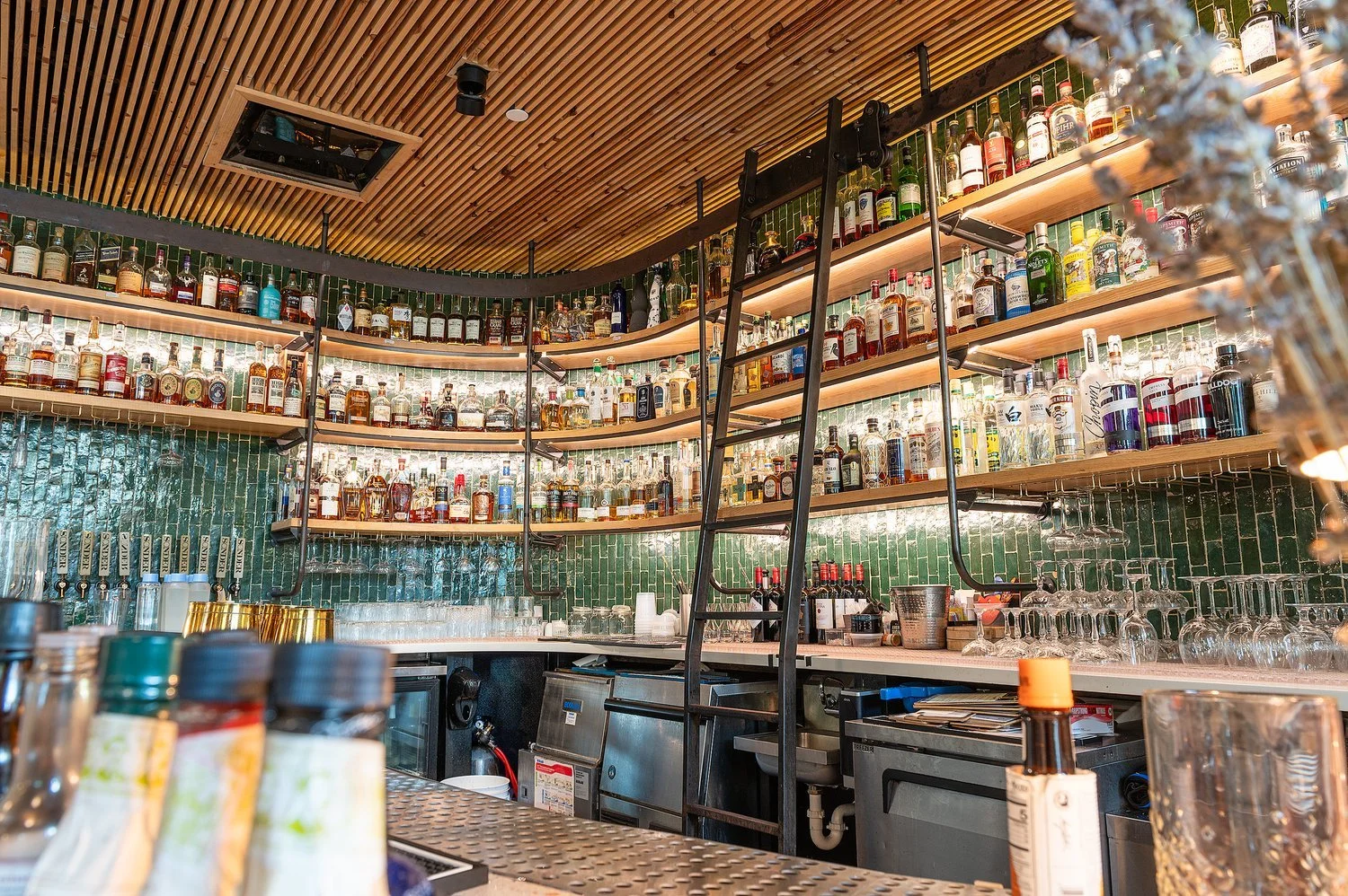 Bar counter with shelves full of various liquor bottles, glasses, and bar tools in a modern, well-lit setting.